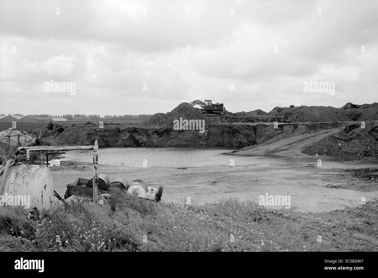 Lagerung giftiger Böden Rijsenhout, Lagerbereiche, Standorte, Rijsenhout, 10-06-1985 Whizgle News, Dutch Desk, Niederlande, 1950-2000 Eine Baustelle weist einen großen Erdhügel im Hintergrund auf, in der Nähe arbeiten schwere Maschinen. Im Vordergrund deuten abgebrannte Fässer und ein wasserdurchtränktes Gebiet auf jüngste Ausgrabungsaktivitäten hin, umgeben von bewölktem Himmel und einem fernen Horizont, der mit weiteren Erdwerken gefüllt ist. Stockfoto