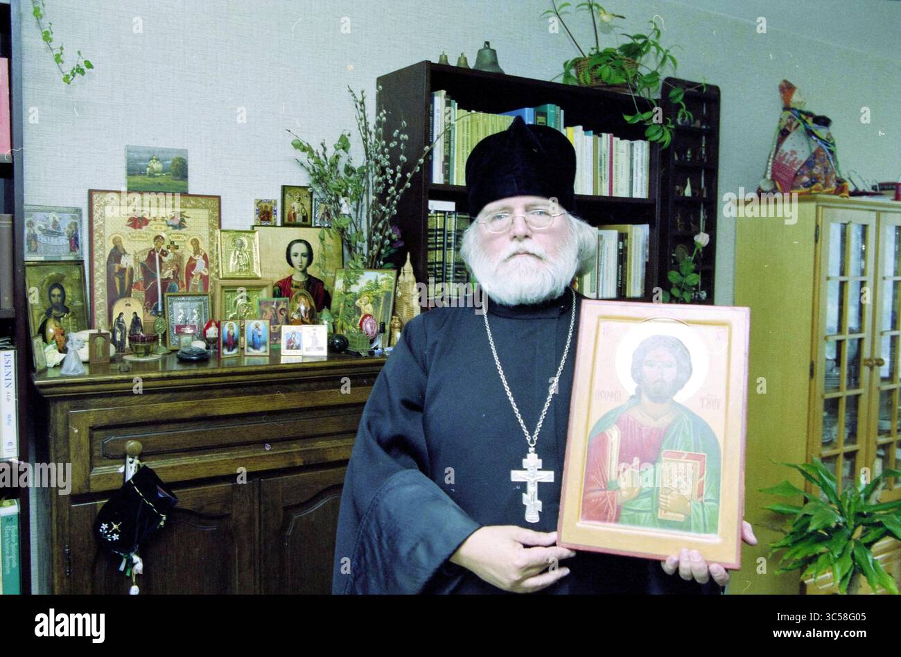 Mr. Messe, Priest Russian Orthodox Church., 30-09-2001 Whizgle News, Dutch Desk, Niederlande, 1950-2000 Ein Mann in traditioneller religiöser Kleidung hält stolz eine Ikone Christi, umgeben von verschiedenen religiösen Artefakten und Bildern auf einem Regal hinter ihm. Das Ambiente bietet eine Mischung aus Büchern und Pflanzen, die eine ruhige Atmosphäre schaffen. Stockfoto