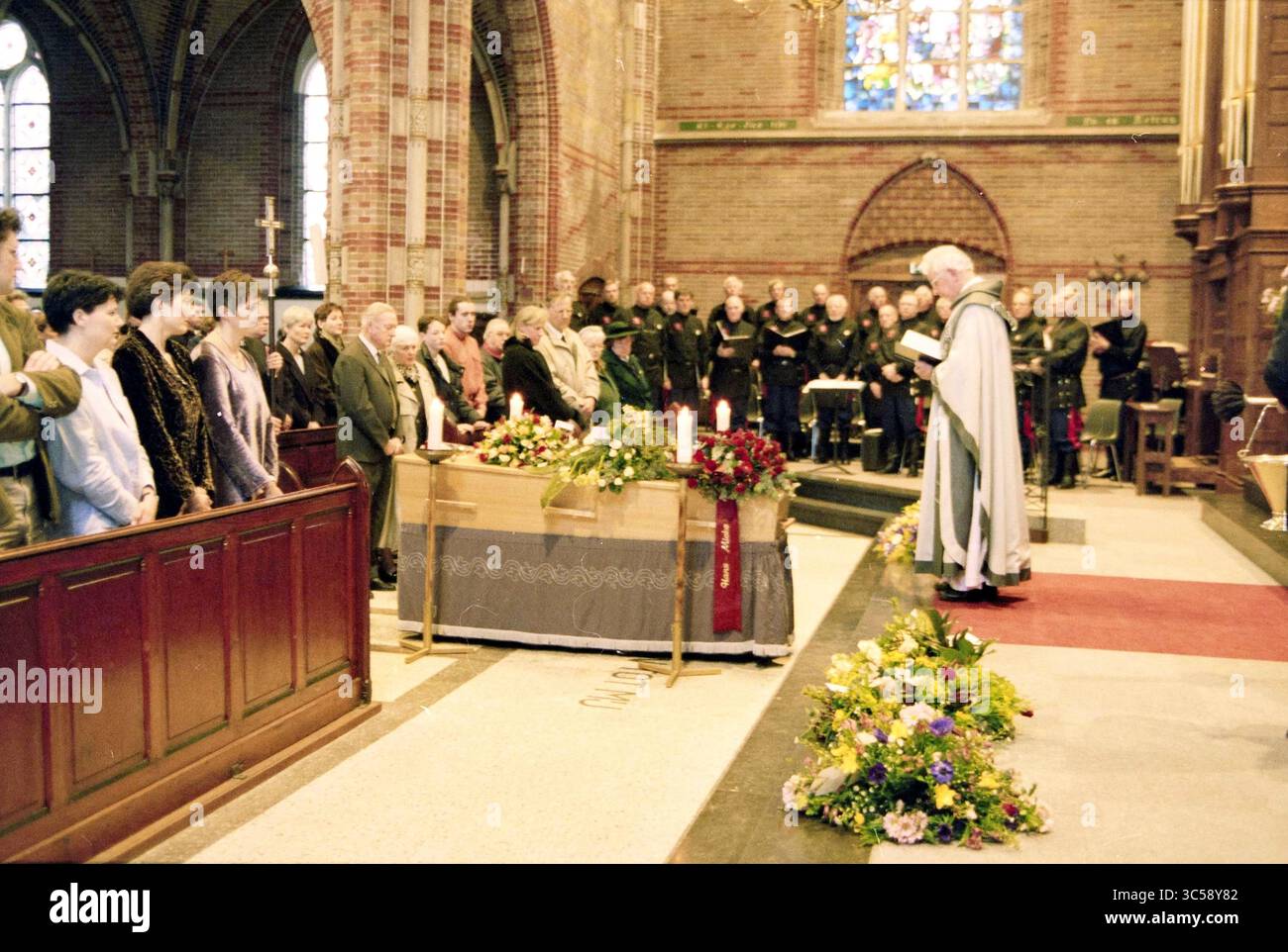 Beerdigung Jan Makkes, 18-01-1999 Whizgle News, Dutch Desk, Niederlande, 1950-2000 in einer großen Kirche findet Eine feierliche Zeremonie statt, an der sich die Teilnehmer zum Gedenken versammeln. An der Vorderseite spricht ein Geistlicher in der Nähe eines mit Blumen geschmückten Schatulls mit herzlichen Worten, während das Publikum aufmerksam zuhört und die düstere Atmosphäre reflektiert. Buntglasfenster erzeugen ein dezentes Leuchten, das die ehrfürchtige Szene unterstreicht. Stockfoto