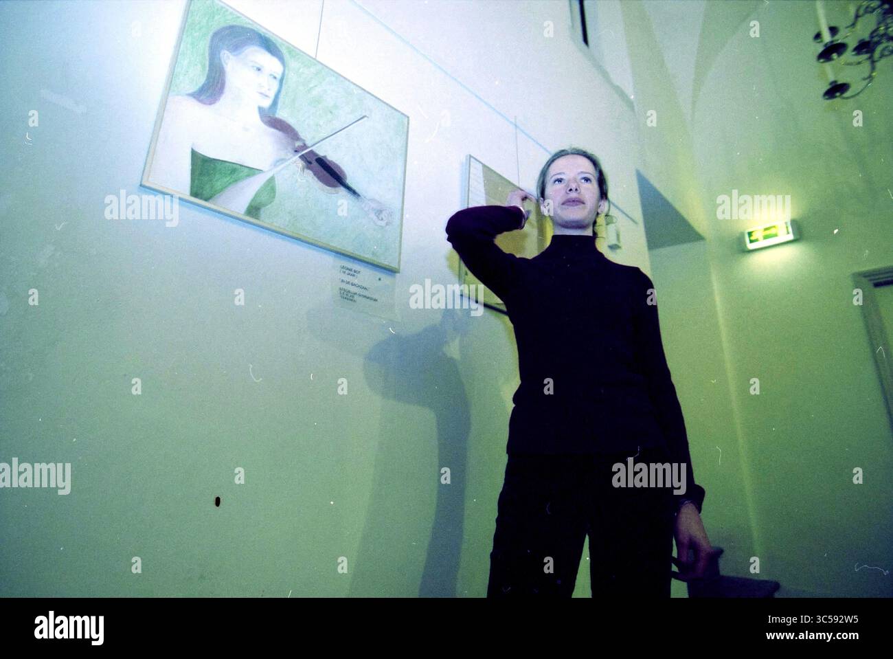 Frau neben dem Gemälde „in the Bach Hall“ Whizgle News, Dutch Desk, Niederlande, 1950-2000 Eine Frau in schwarzem Outfit steht selbstbewusst, mit einer Hand hochgehoben und die andere auf ihrer Hüfte, während sie neben einem Kunstwerk einer Geige spielenden Frau posiert. Das Ambiente verfügt über eine sanfte Beleuchtung, die die künstlerische Atmosphäre unterstreicht. Stockfoto