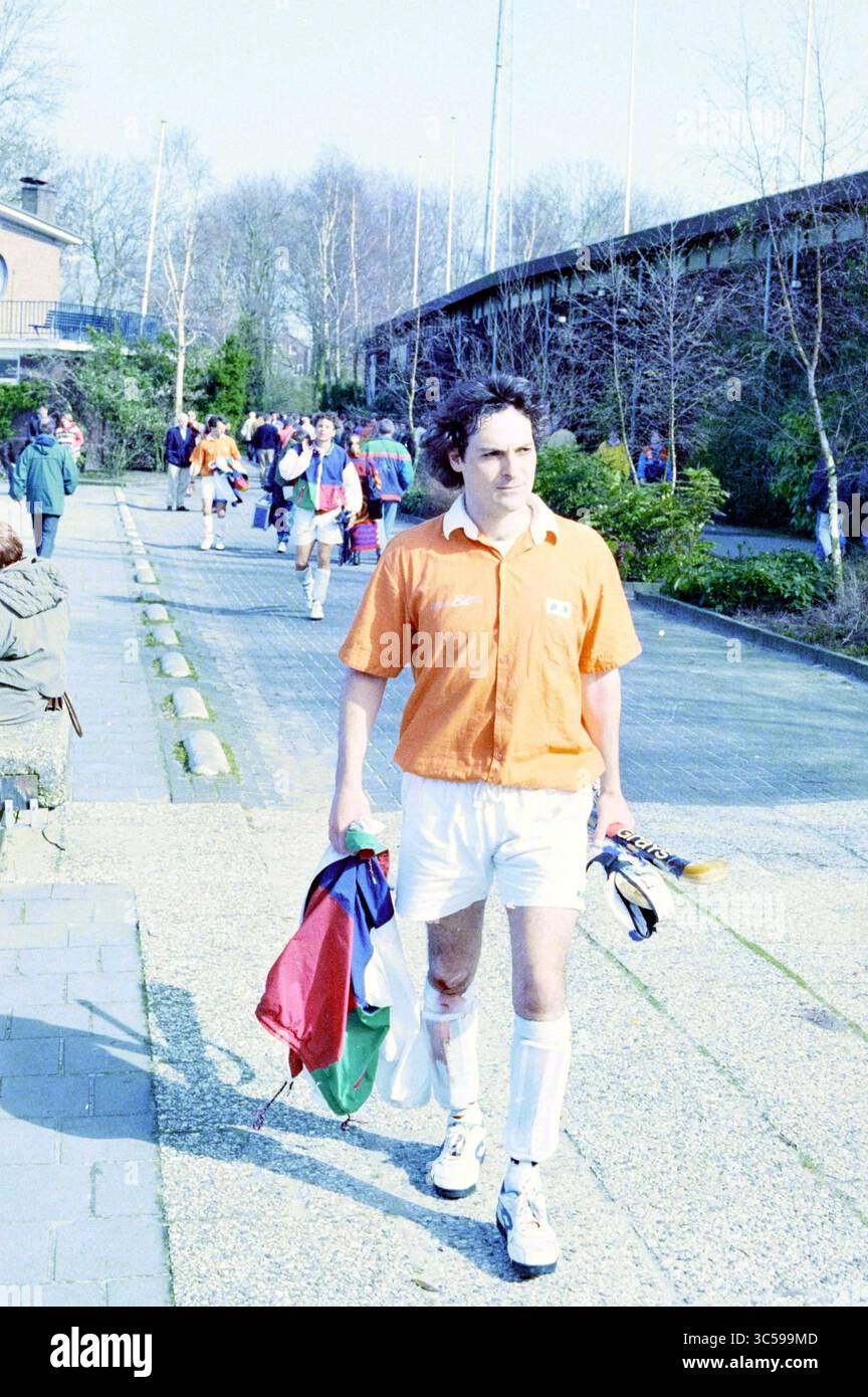 Hockeyspiel Bloemendaal-Amsterdam, 01-04-1995 Whizgle News, Dutch Desk, Niederlande, 1950-2000 Ein Spieler in einem orangen Trikot geht selbstbewusst den Weg entlang, hält Sportausrüstung und trägt weiße Shorts, während eine Gruppe von Athleten im Hintergrund auf eine Sportanlage zusteuert. Die Umgebung ist mit Bäumen und einem gepflasterten Fußweg ausgestattet, der eine ungezwungene und dennoch fokussierte Atmosphäre widerspiegelt. Stockfoto