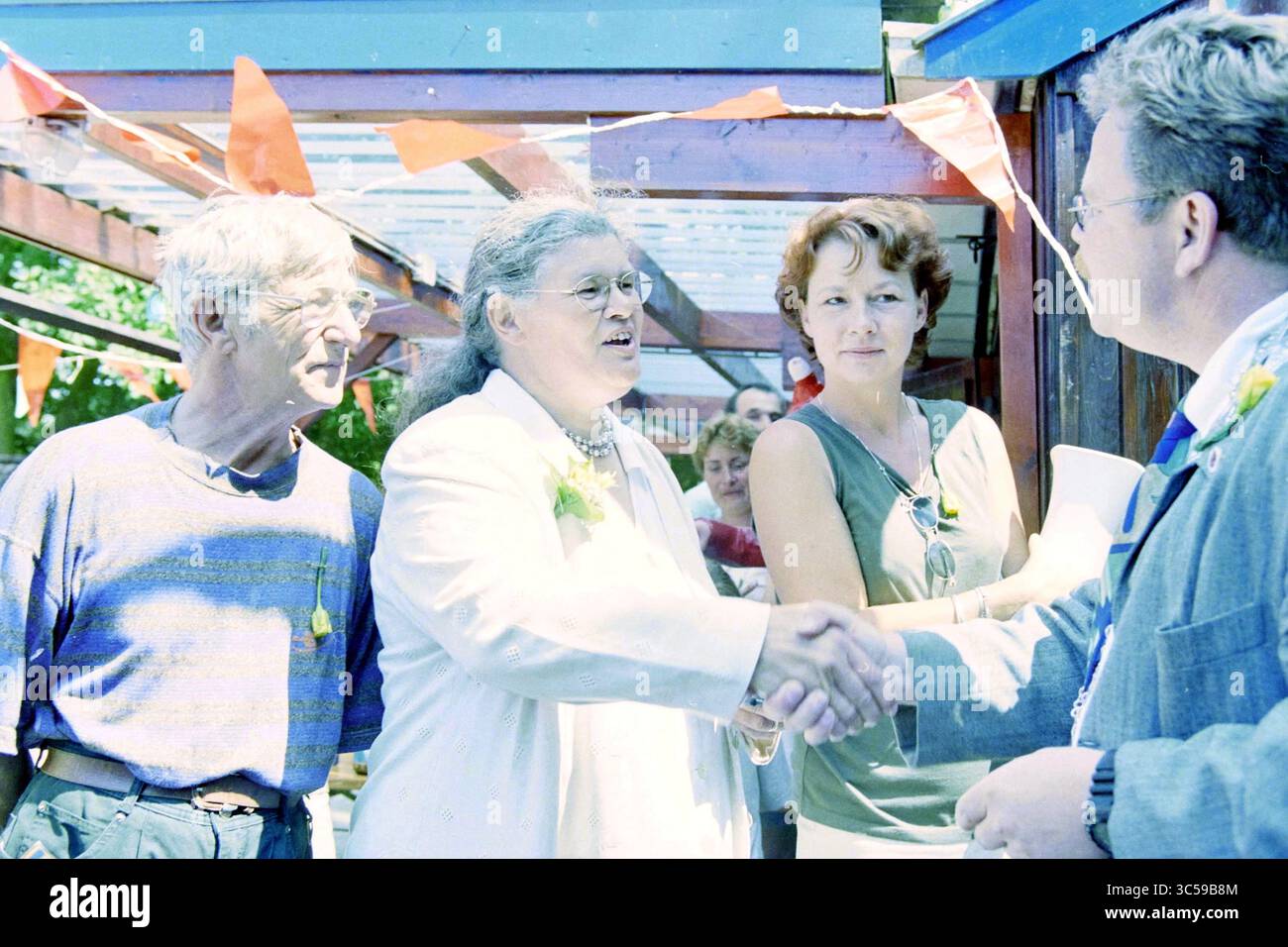 Tante Eef, Medaille, Santpoort-Noord, Santpoort-Noord, 23-07-1999 Whizgle News, Dutch Desk, Niederlande, 1950-2000 Eine Gruppe von Menschen versammelt sich im Freien, um ein freundliches Gespräch zu führen. Eine Frau in einem weißen Outfit schüttelt einer anderen Person warm die Hände, während andere zuschauen, was ein Gefühl von Kameradschaft schafft. Farbenfrohe Dekorationen und ein zwangloses Ambiente lassen eine festliche Atmosphäre vermuten. Stockfoto