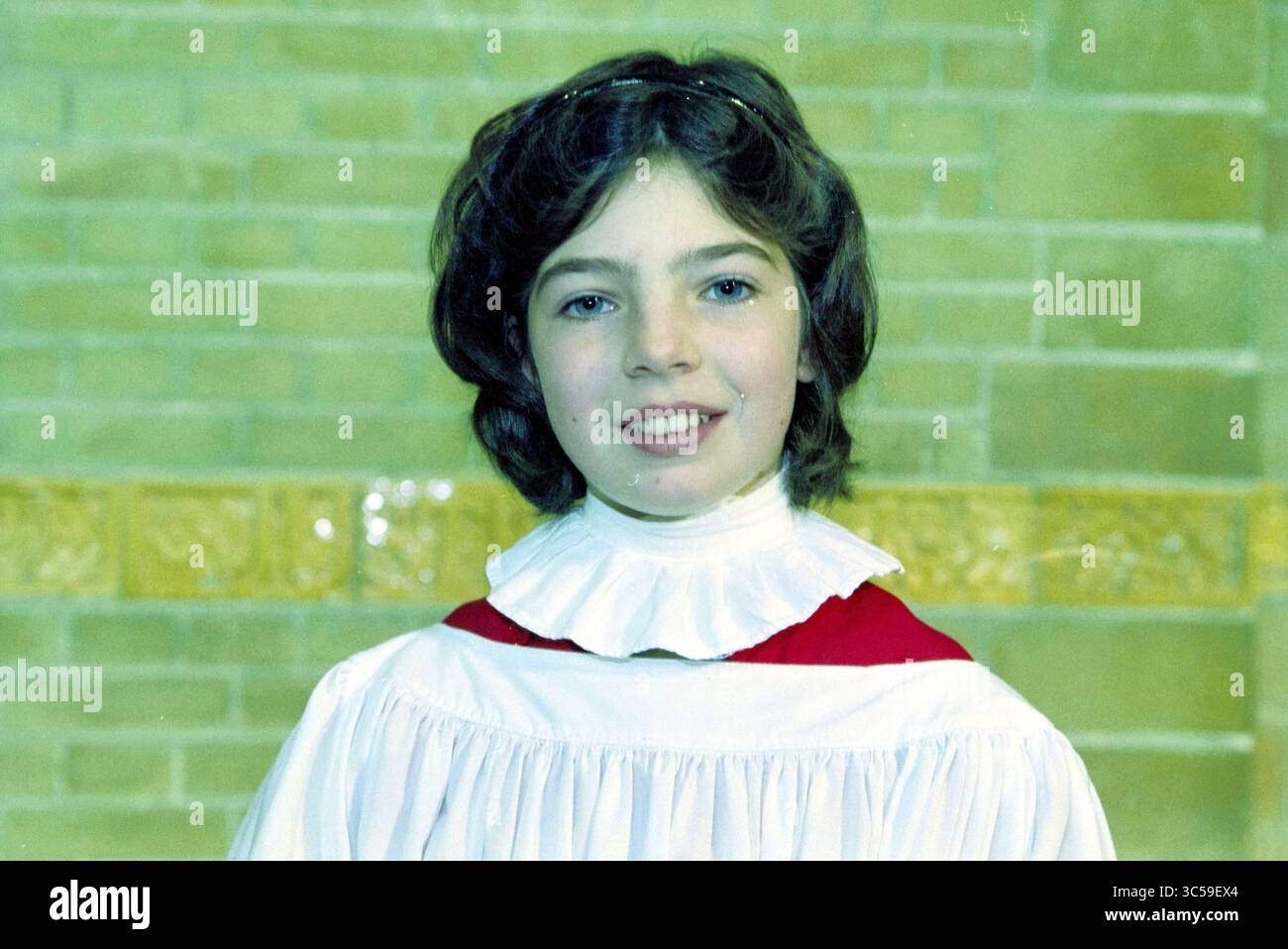 Porträts der Mitglieder der Haarlem Chorschule, 13-10-2001 Whizgle News, Dutch Desk, Niederlande, 1950-2000 Ein junger Chormitglied lächelt selbstbewusst, gekleidet in einem traditionellen Chormantel mit Rüschenkragen und einem leuchtenden roten Akzent. Der Hintergrund ist mit strukturierten Ziegeln versehen, die eine warme und einladende Atmosphäre schaffen. Stockfoto