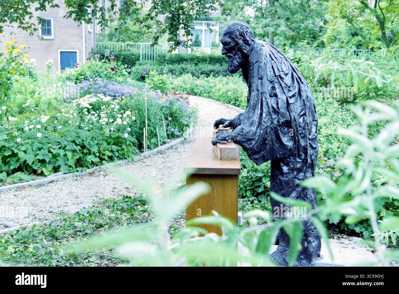 Joseph Statue, Mr. Mesman, Hoofddorp, Niederlande, 01-10-2003 Whizgle News, Dutch Desk, Niederlande, 1950-2000 Eine verwitterte Statue eines älteren Mannes lehnt sich nachdenklich über ein hölzernes Geländer, umgeben von einem üppigen Garten mit lebhaften Blumen und Grün. Die beschauliche Szene lädt zur Reflexion und Besinnung inmitten der Schönheit der Natur ein. Stockfoto