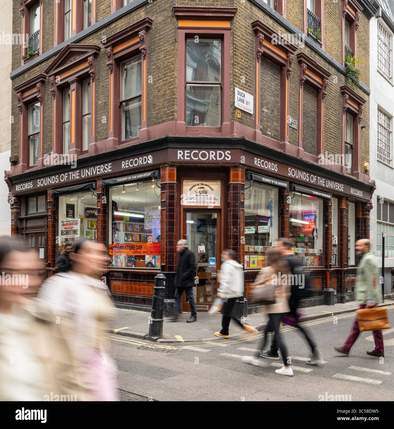 Fußgänger laufen an einer belebten Straßenecke in Soho, London, vorbei an den legendären Sounds des Universe Records Stores, und Bewegungsunschärfen sorgen für ein dynamisches Gefühl. Stockfoto