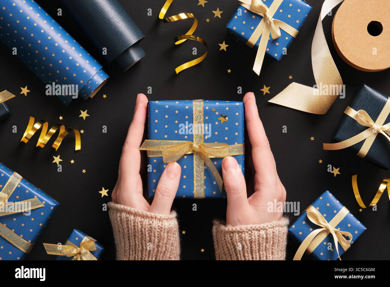 Die Hände halten eine blaue Geschenkbox mit goldenem Band, umgeben von Geschenkverpackungen auf dunklem Hintergrund Stockfoto