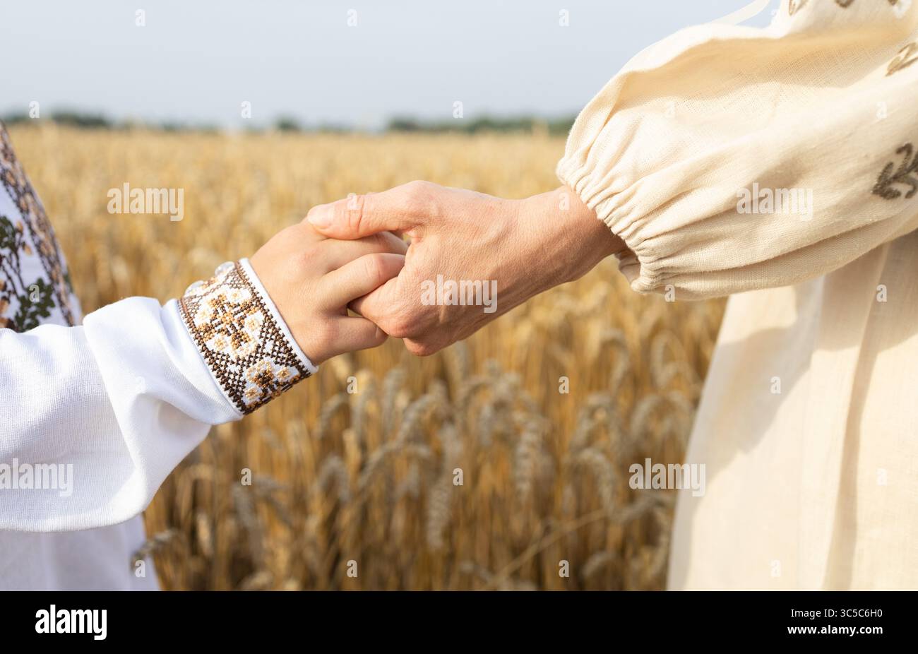Frauen- und Kinderhand in einem bestickten Hemd halten sich vor dem Hintergrund des Weizenfeldes. Ukrainische Familie, Einheit, Fürsorge, Gefühl der Unterstützung und Stockfoto