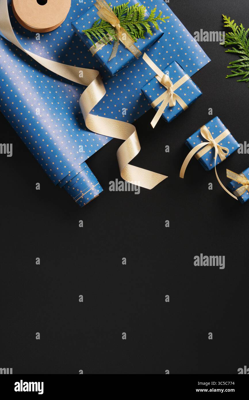 Flache Komposition mit blauen Geschenken mit goldenen Bändern, braunen Papieranhänger, grünen Thuja-Zweigen auf schwarzem Hintergrund. Platz für Ihren Text. Stockfoto