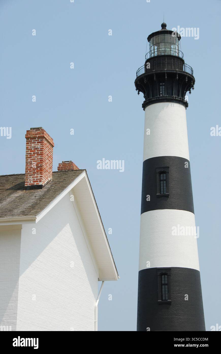 Ein schwarz-weiß gestreifter Leuchtturm mit einem angrenzenden weißen Haus und roten Schornsteinen vor einem klaren blauen Himmel. Eine klassische Küstenszene. Stockfoto