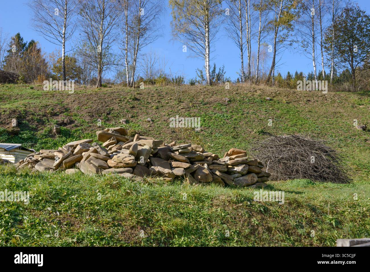 Großabhang des Hinterhofhügels mit Haufen flacher Steine und Reisig auf grünem Gras in Skhidnytsia, Westukraine. Stockfoto
