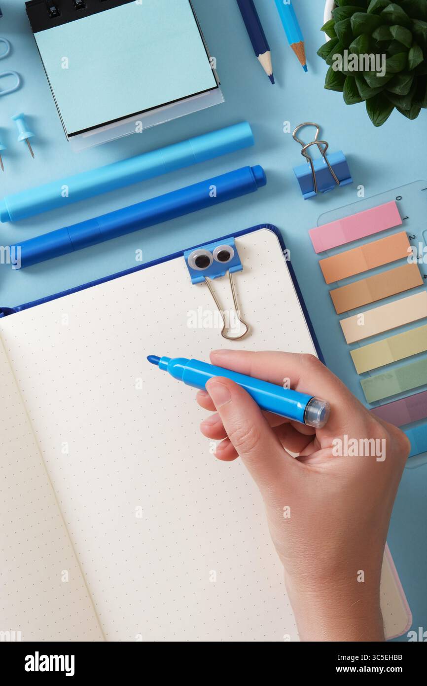 Die Hand hält einen blauen Filzstift vor einem geöffneten Notizbuch mit leerer Seite. Um blaue Büroartikel Stifte, Bleistifte, Filzstifte, s Stockfoto