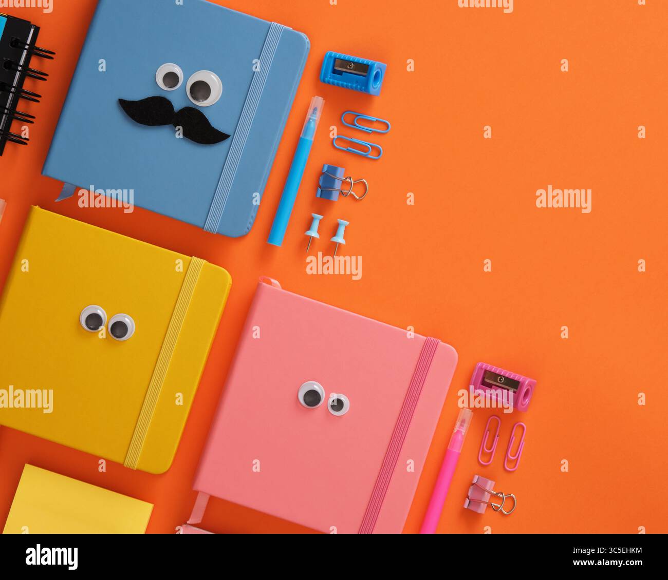 Bunte Notizbücher mit Googly Eyes und lustige Büromaterialien auf orangefarbenem Hintergrund. Kopierraum. Stockfoto