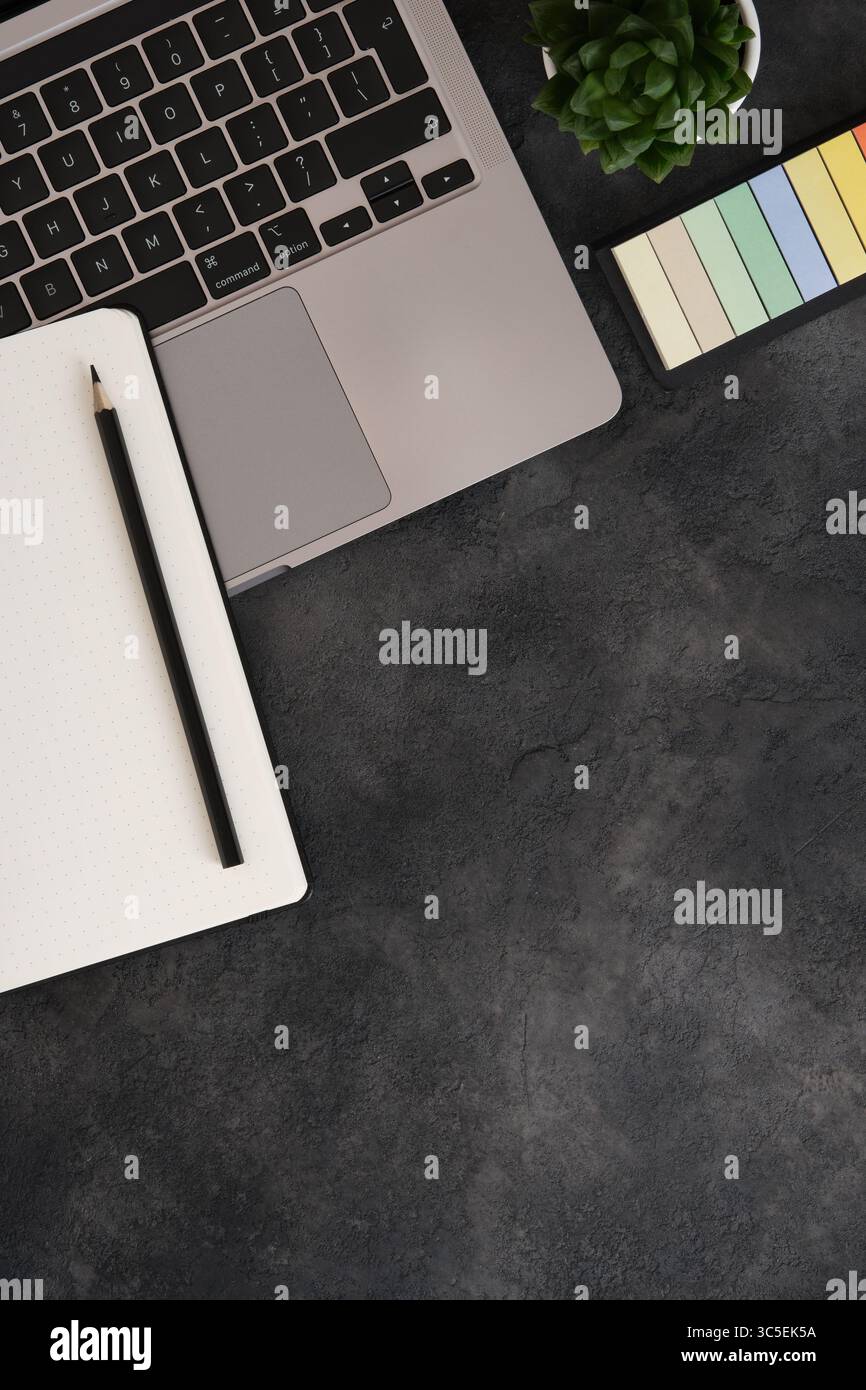 Minimalistischer Arbeitsbereich mit Laptop, Kugelplaner, Stift, Aufklebern und saftiger Pflanze auf dunklem, strukturiertem Hintergrund. Kopierbereich. Stockfoto
