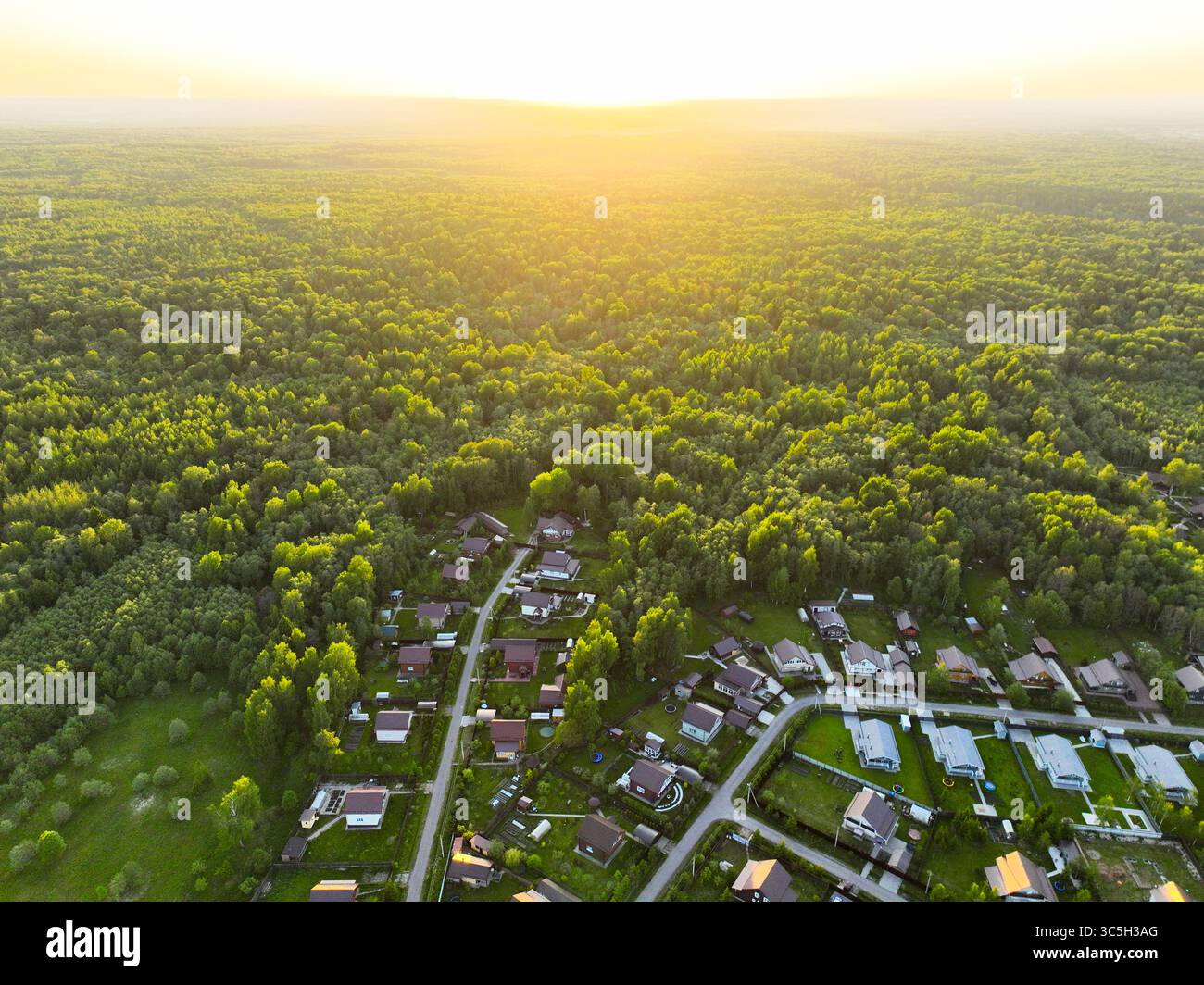 Ein Luftbild fängt den warmen Sonnenuntergang über einem weitläufigen grünen Wald mit einem ländlichen Dorf im Vordergrund ein Stockfoto