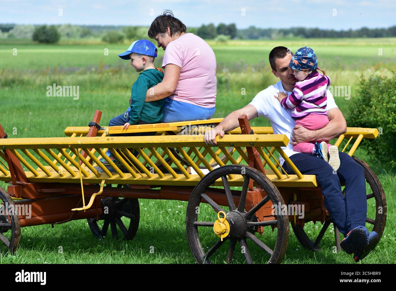 Die Familie genießt eine Fahrt mit einem Holzwagen durch das grüne Feld. Mehrgenerationsbindung und Outdoor-Erholung in ländlichen Umgebungen. Stockfoto