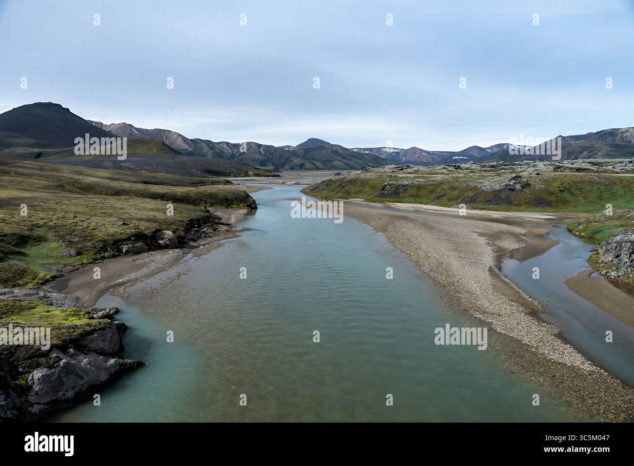 Blick auf einen Gletscherfluss, der sich durch eine raue Landschaft aus grünem Moos und schwarzem vulkanischem Gestein unter einem riesigen, bewölkten Himmel schlängelt, Kirkjubaejarklaustur, Skaftárhreppur, Island. Stockfoto