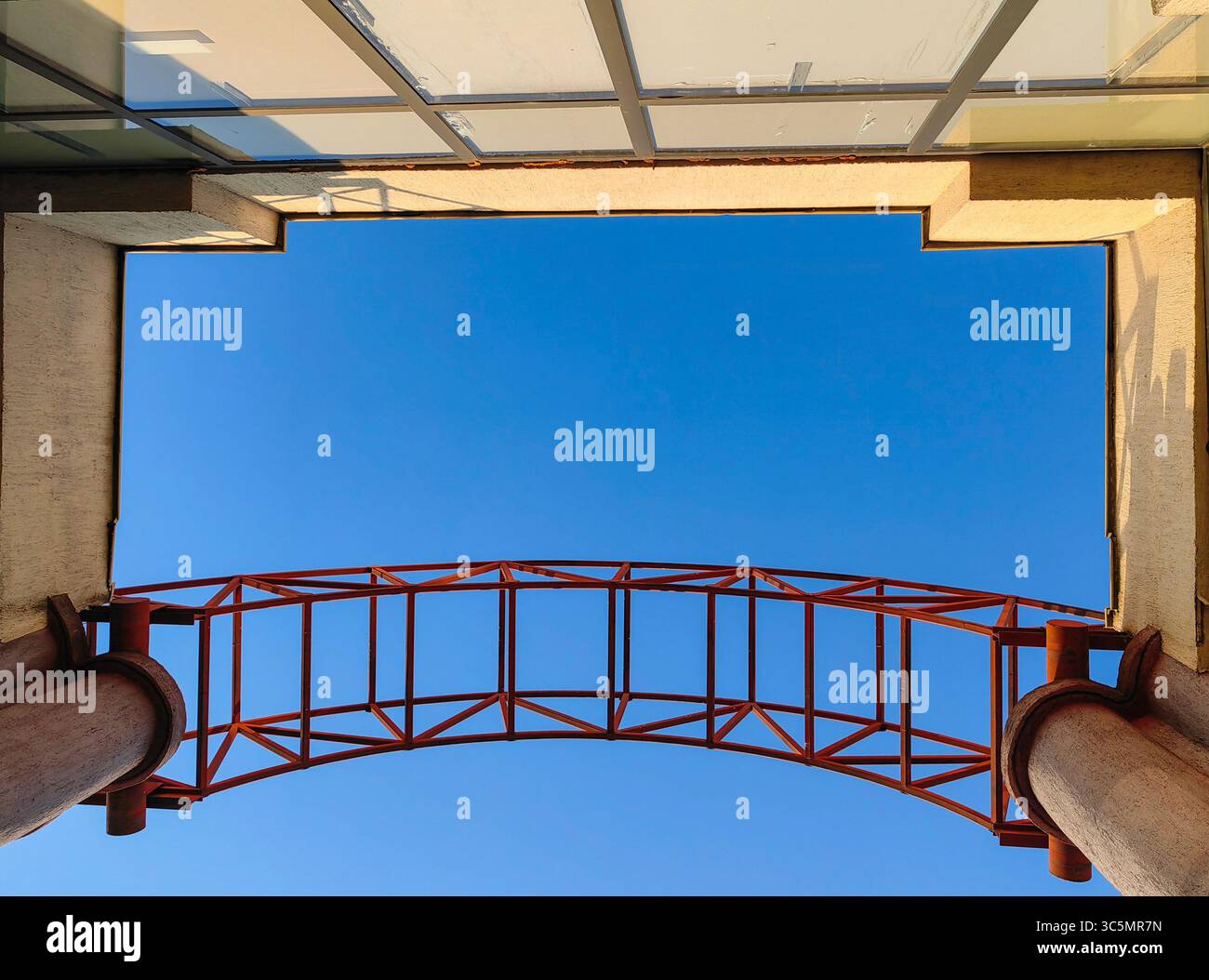 Blick nach oben: Roter Metallbogen und Betongeometrie gegen den blauen Himmel Stockfoto