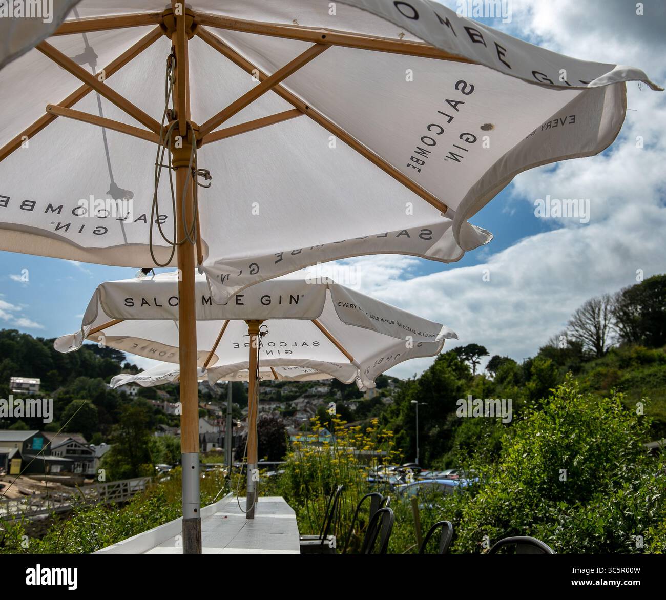Sitzbereich im Freien mit Sonnenschirmen der Marke Salcombe Gin mit Blick auf einen malerischen Hügel und den bewölkten Himmel von Salcombe Devon Stockfoto