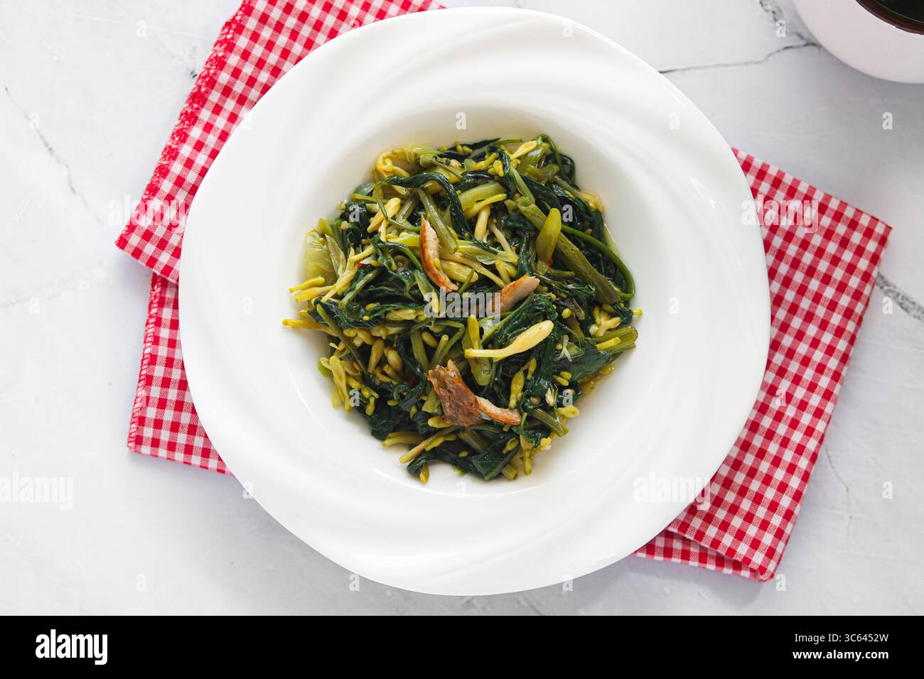 Ein Teller mit gebratenem Gemüse im Manado-Stil (Kangkung Bunga Pepaya), auf dem frische, belaubte Zutaten und aromatische Gewürze präsentiert werden. Stockfoto