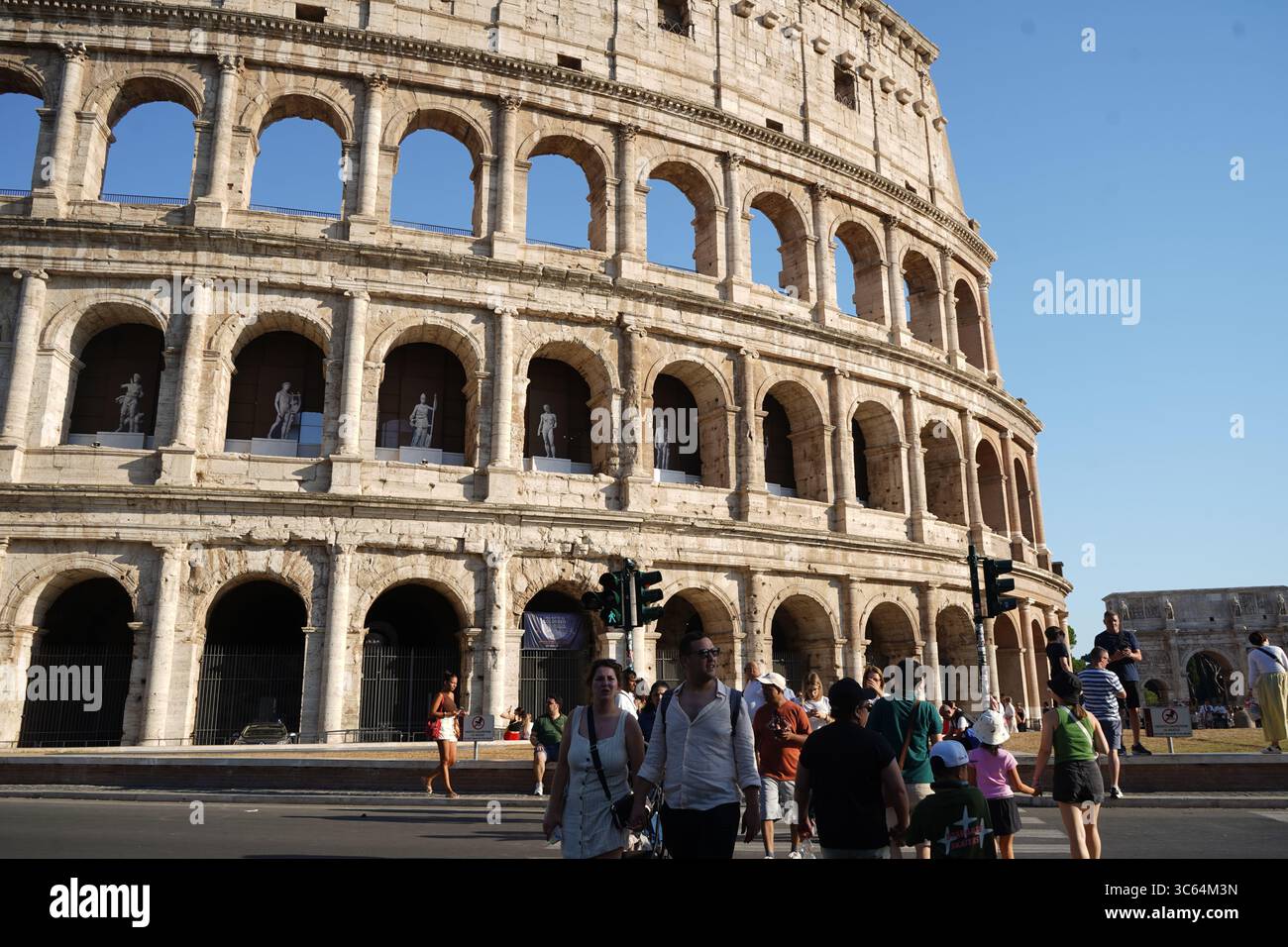 Touristen spazieren vorbei am imposanten Kolosseum, einem antiken römischen Amphitheater. Seine großen Bögen und seine historische Präsenz prägen die sonnige Stadtlandschaft. Stockfoto