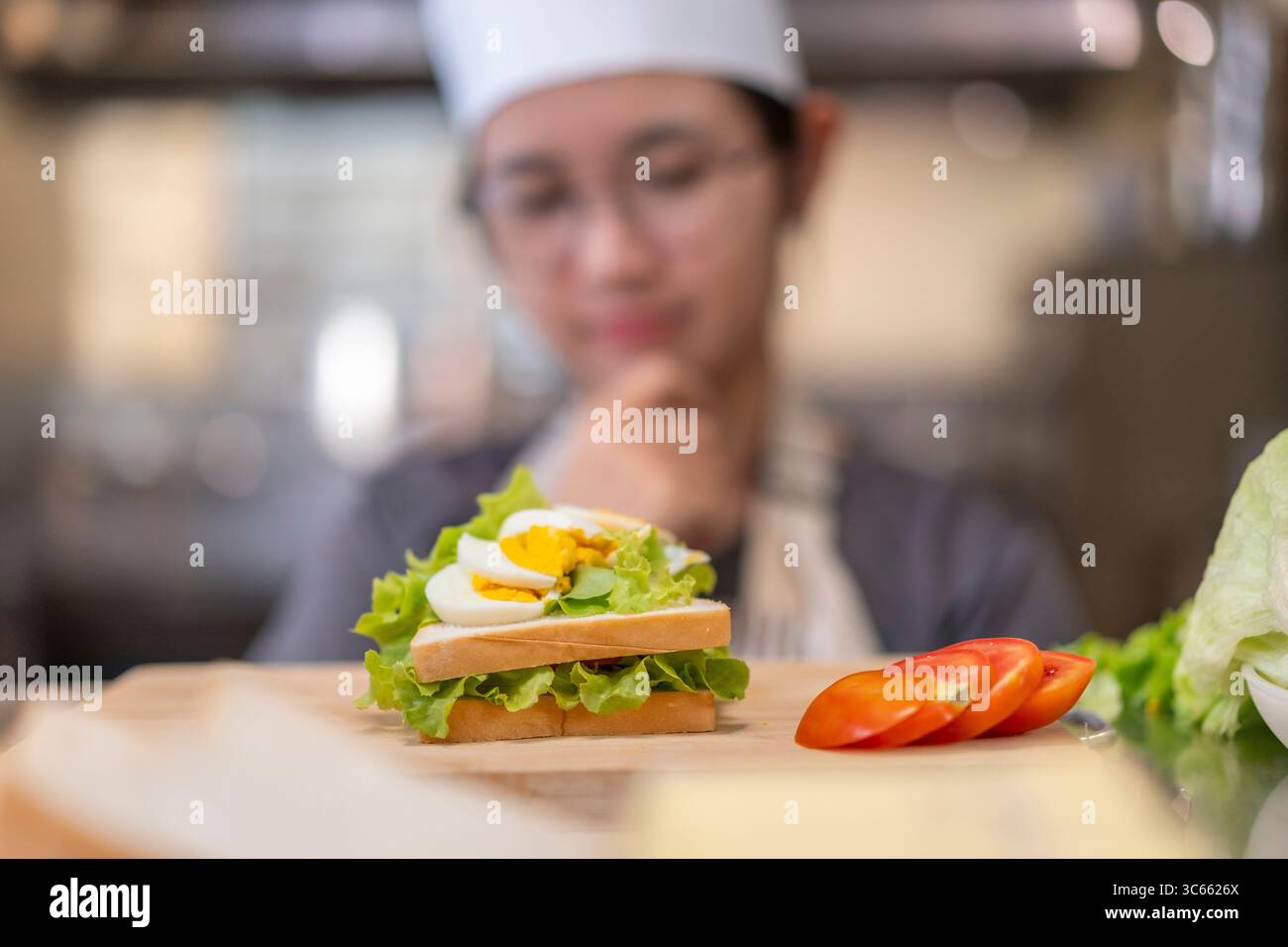 Eine junge Küchenchefin in Uniform untersucht sorgfältig ein von ihr zubereitetes Sandwich und zeigt Fokus, Kreativität und kulinarische Fähigkeiten in einer kommerziellen Küche Stockfoto