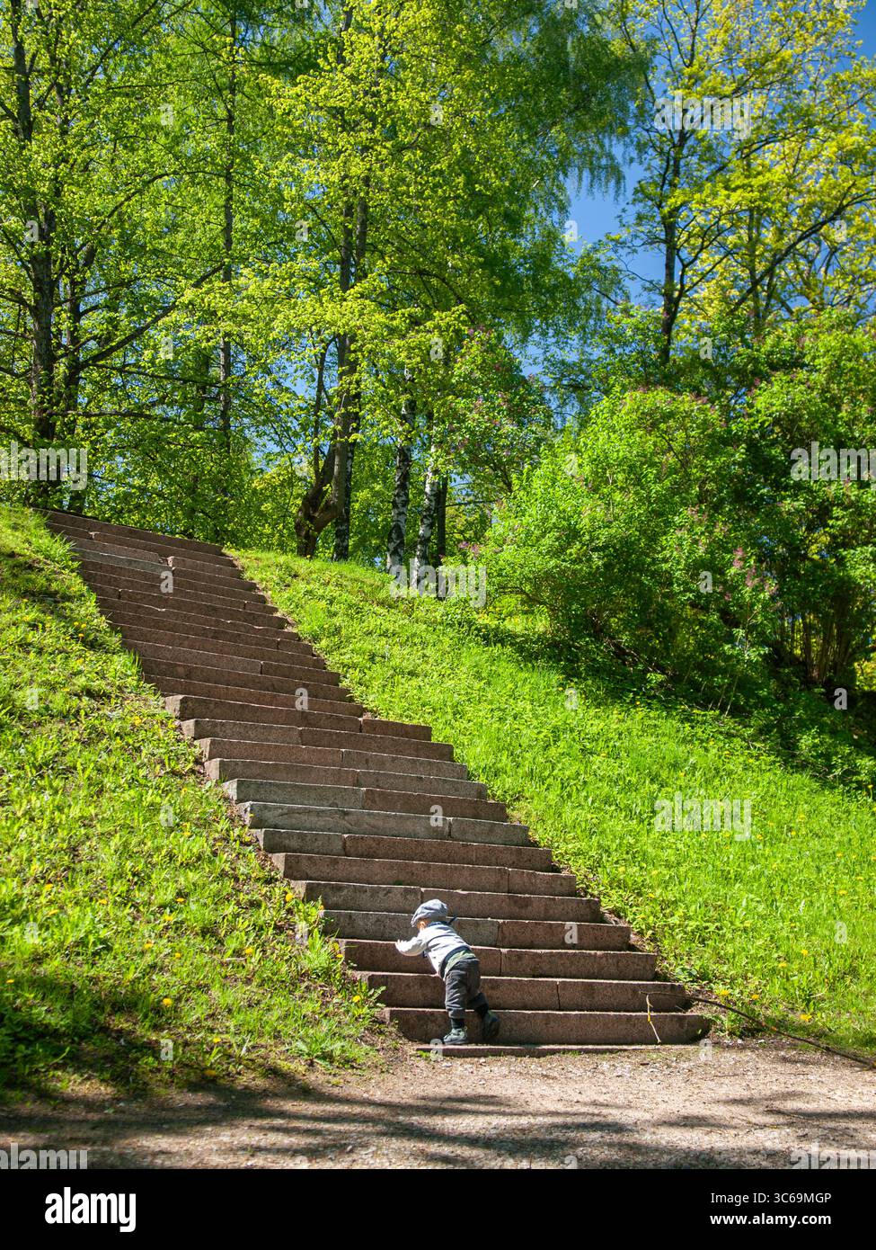 Ein kleines Kind am Fuß einer Steintreppe in einem üppig grünen Park, der den Beginn einer Reise mit frischem Frühlingslaub rund um die Uhr darstellt. Stockfoto
