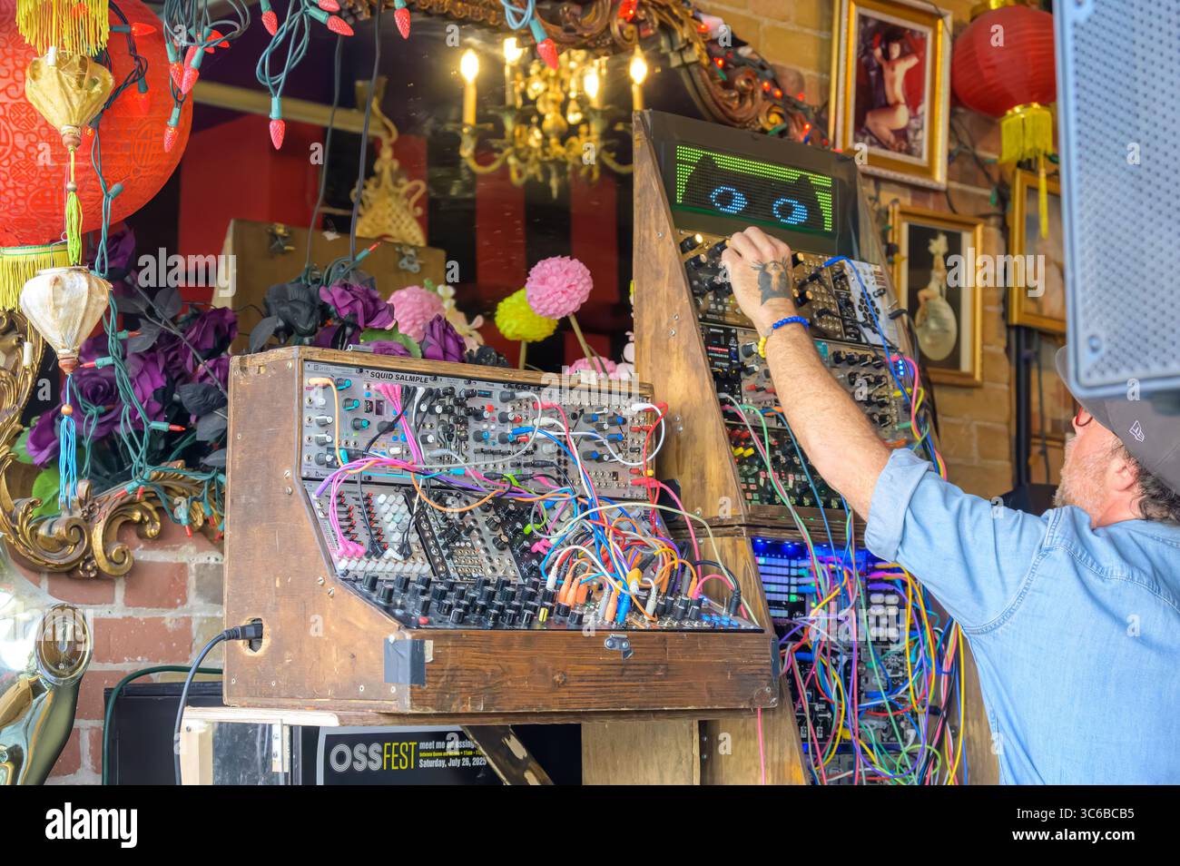 Ein Mann passt während des OSSFest einen modularen Synthesizer an, der von bunten Kabeln, Vintage-Dekor und einem digitalen Katzengesicht umgeben ist. Stockfoto