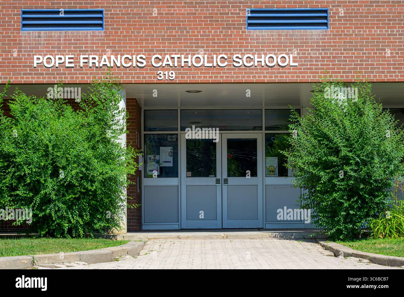 Der Eingang der Papst-Franziskus-katholischen Schule hat eine rote Backsteinfassade, weiße Schriften und Glastüren, die von grünen Sträuchern eingerahmt werden. Stockfoto