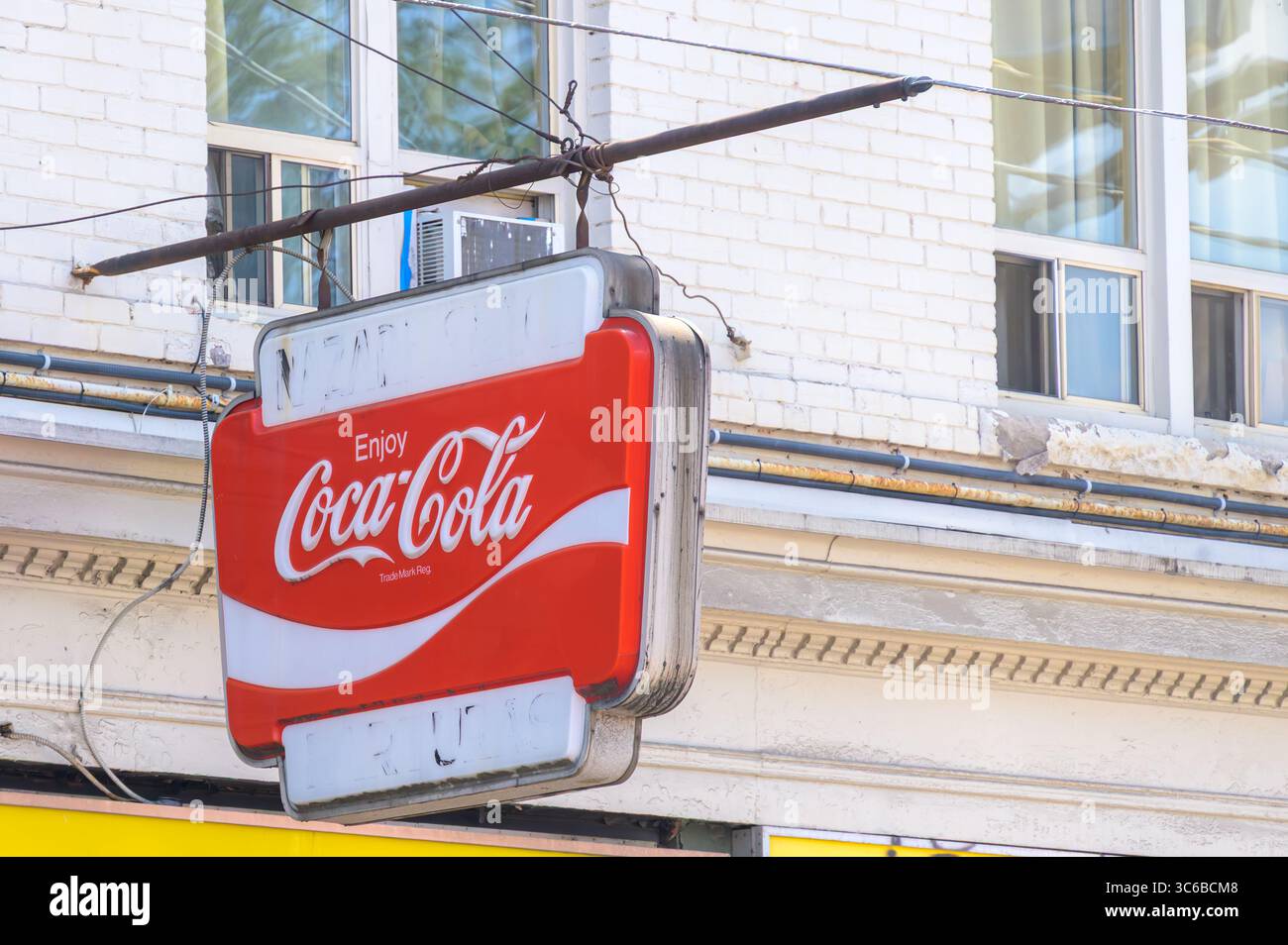 Ein altes rot-weißes Coca-Cola-Schild hängt an einer verwitterten Stange an einem weißen Backsteingebäude mit alten Fenstern darüber. Stockfoto