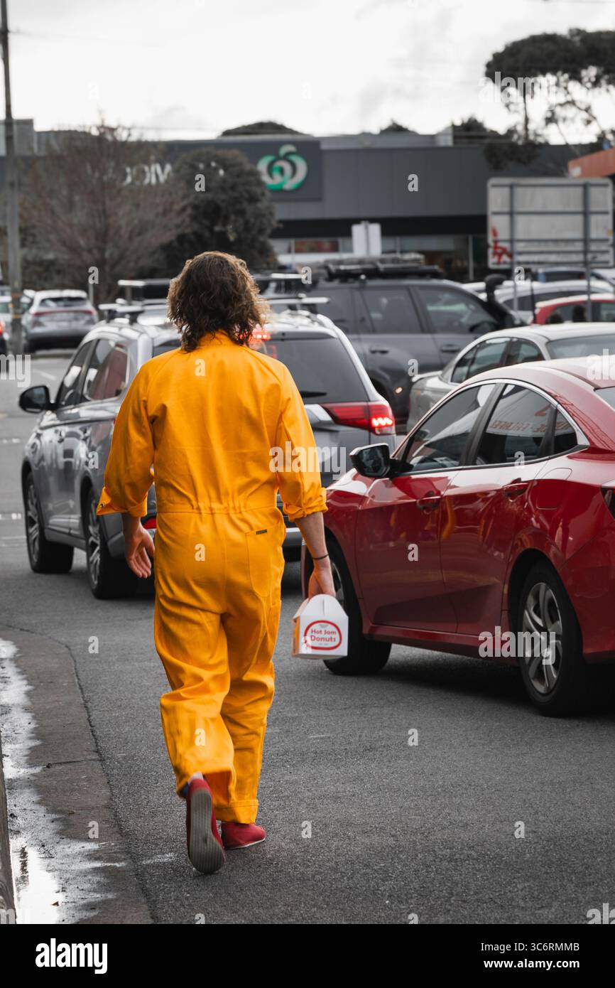 Ein Mann, der mitten auf der Straße läuft, voll orange Outfit, lange Haare, rote Schuhe, rote Autos Stockfoto