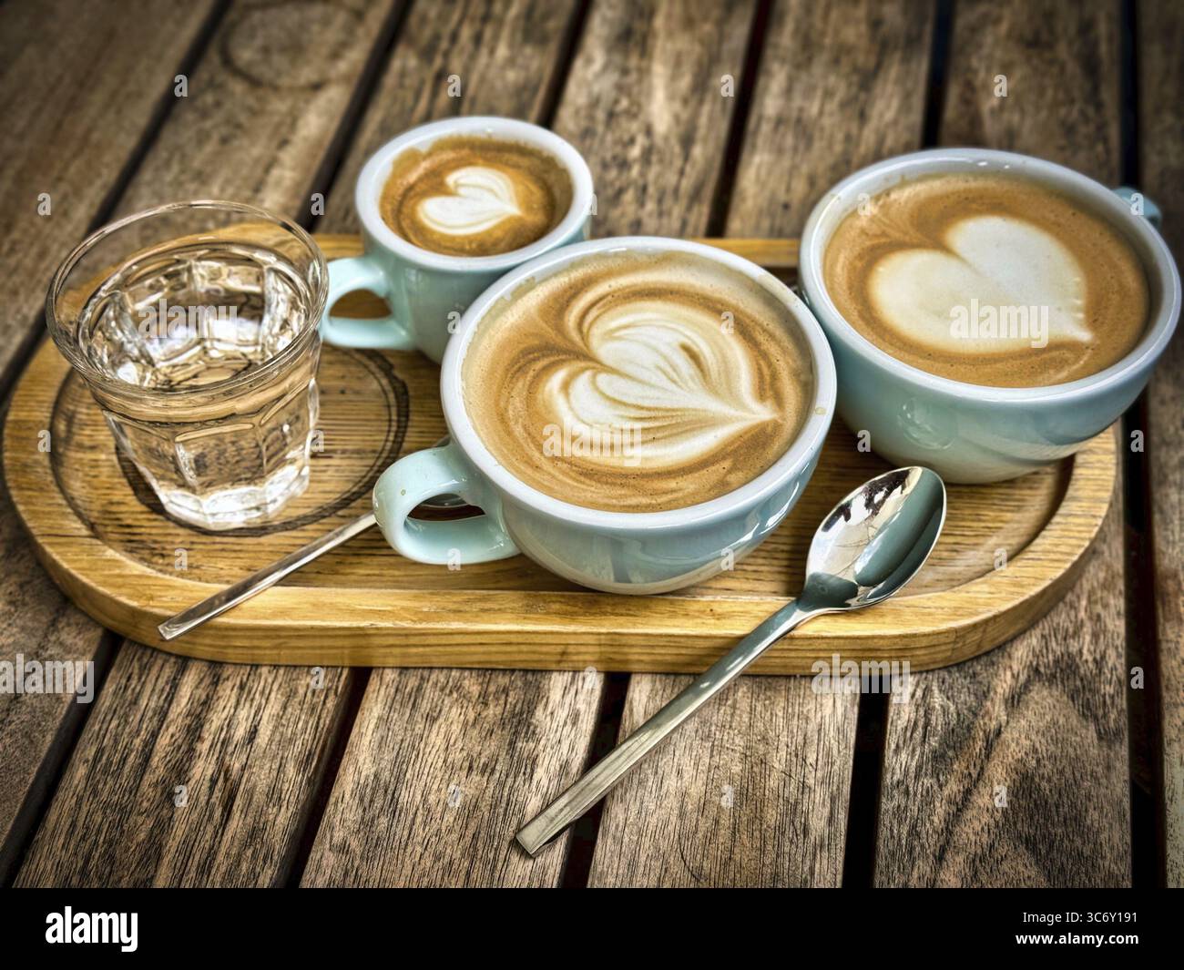 Drei Kaffeetassen mit Herzmuster auf einem Holztablett, begleitet von einem Glas Wasser Stockfoto