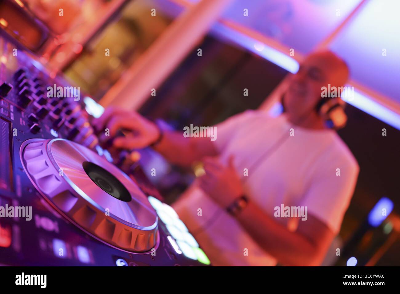 Verschwommener DJ mit Kopfhörern an einem Mischpult mit Plattenspieler und Bedienelementen macht Musik bei einer Veranstaltung in stimmungsvoller Partyatmosphäre mit farbenfrohem Licht Stockfoto