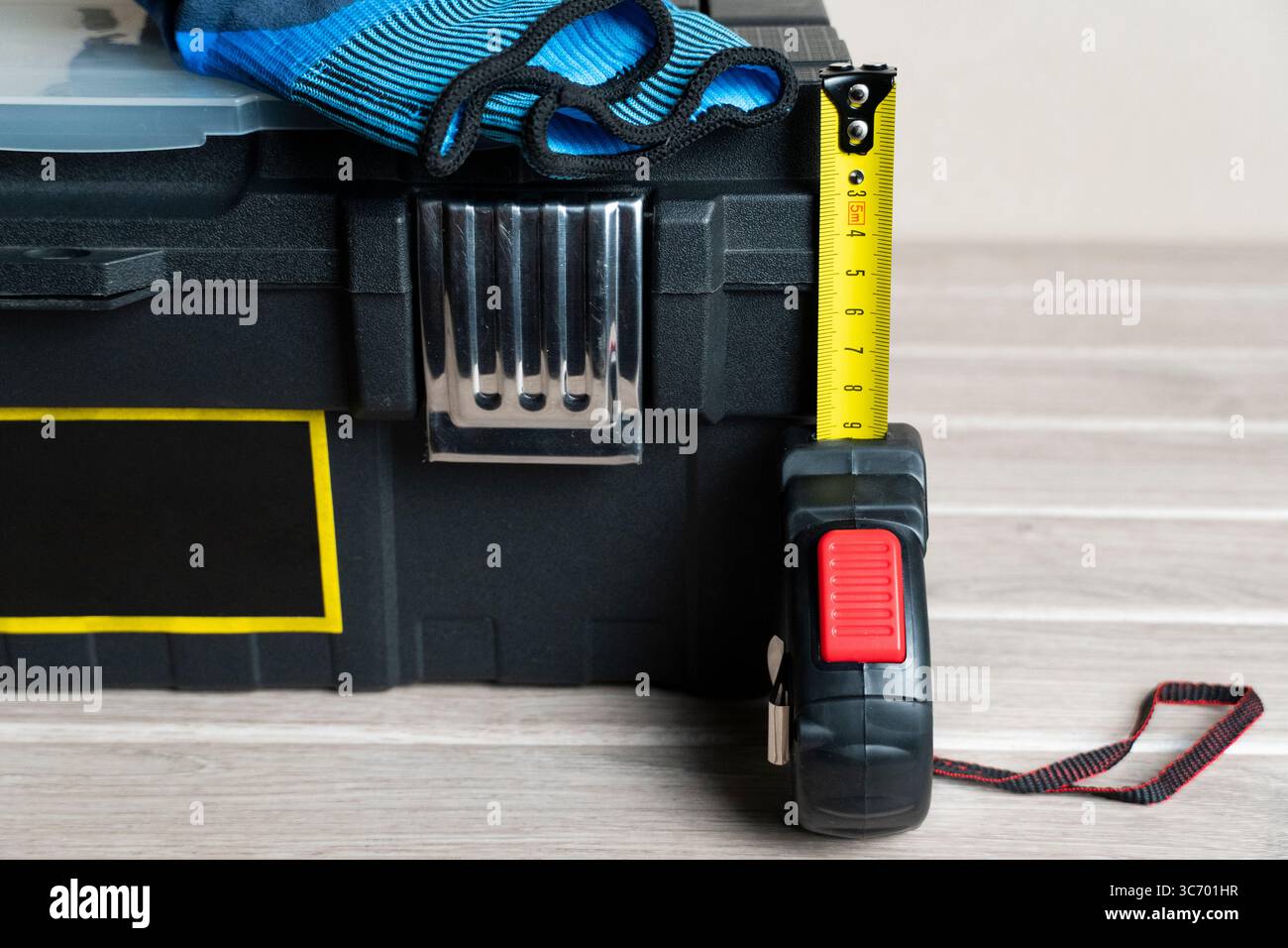 Ein schwarzer Werkzeugkasten mit blauen und schwarzen Arbeitshandschuhen und einem verlängerten gelben Maßband, bereit für Heimwerkerprojekte und Hausreparaturen Stockfoto