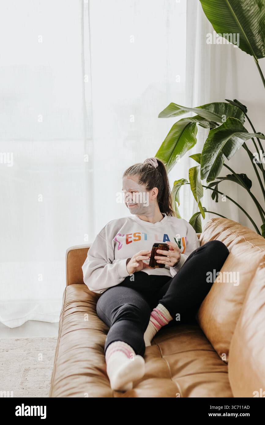 Frau mittleren Alters entspannt sich auf der Couch, während sie am Telefon eine Tonmaske trägt Stockfoto