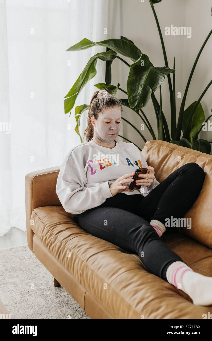 Frau mittleren Alters entspannt sich auf der Couch, während sie am Telefon eine Tonmaske trägt Stockfoto