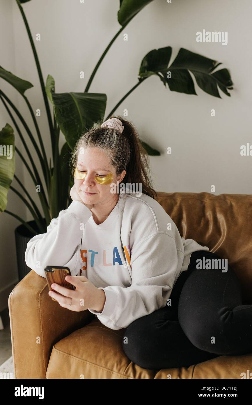 Frau sitzt auf der Couch mit Augenmasken, während sie das Handy benutzt Stockfoto