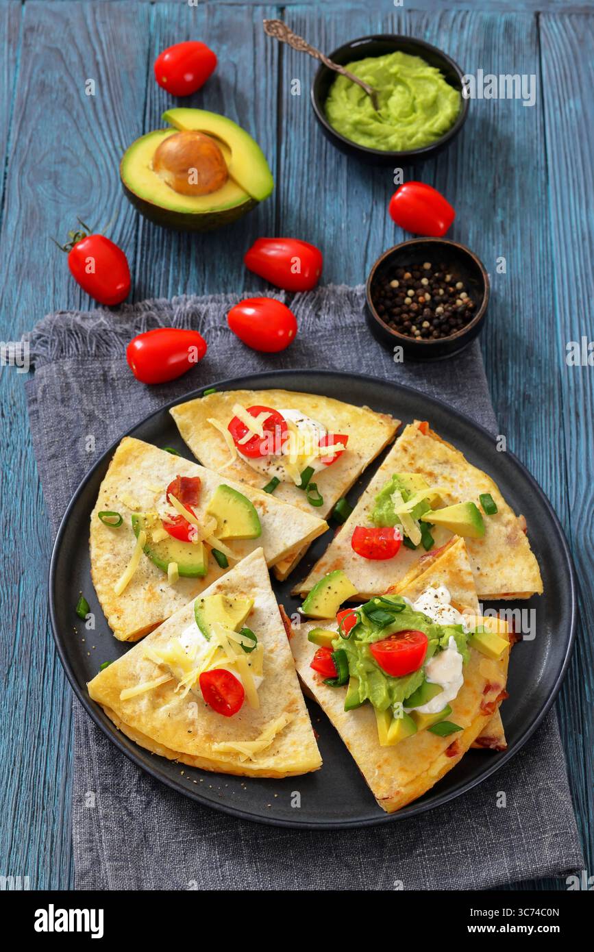 Quesadilla mit Käse und gebratenem Speck, garniert mit Sauerrahm, Guacamole, Avocado und Tomaten auf einem Teller auf Holztisch mit Zutaten, vertikal Stockfoto