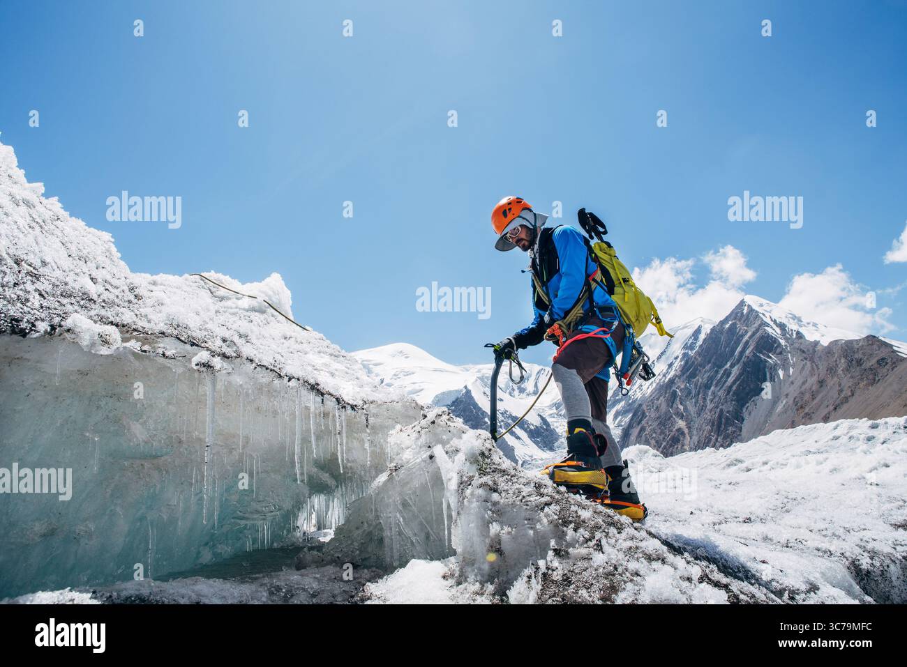 Bergsteiger in pulsierenden Gewerken überqueren Gletscherspalten während des Lenin Gipfels unter klarem Himmel mit schneebedeckten Gipfeln im Hintergrund. Extreme Akti Stockfoto
