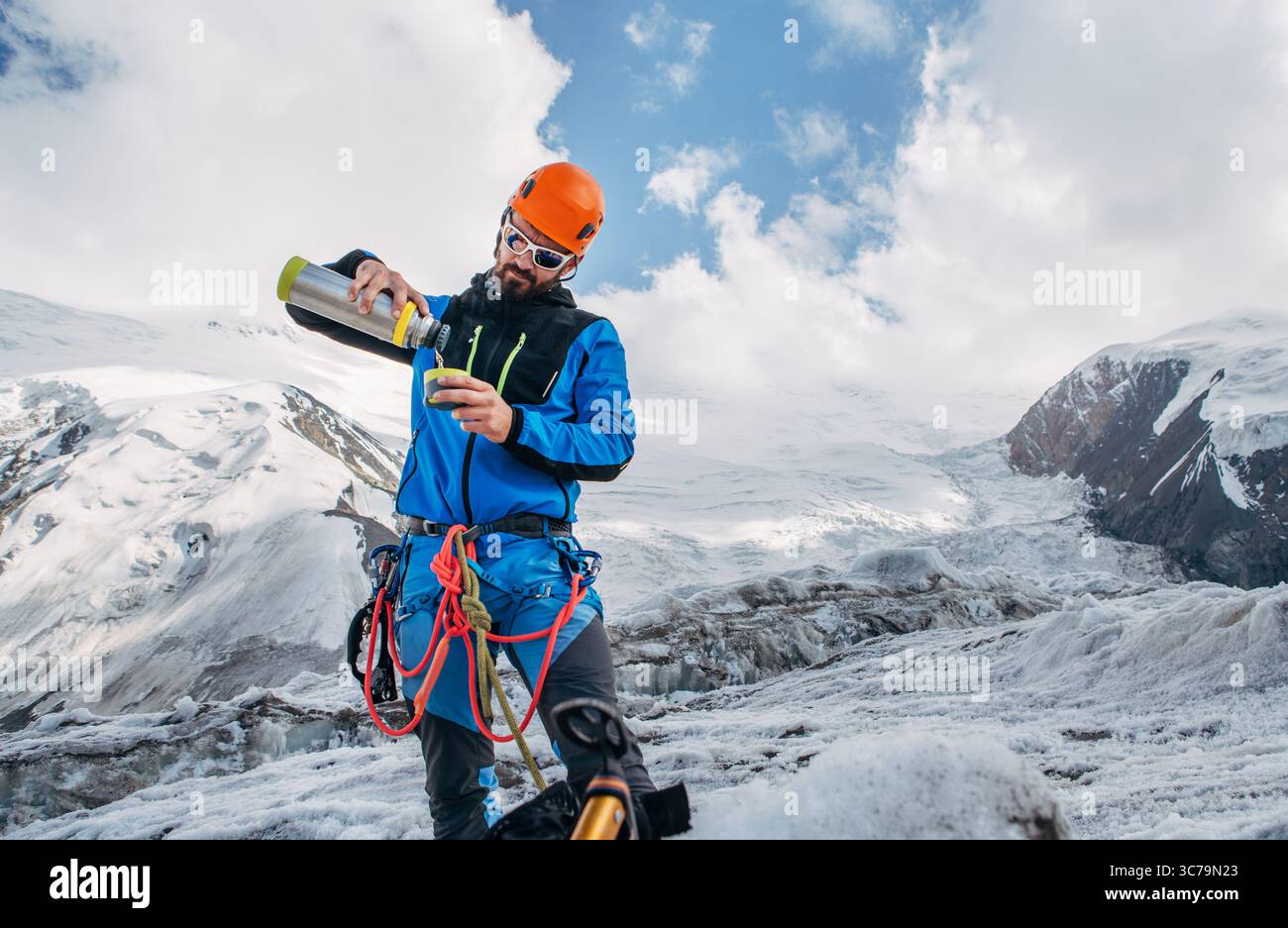 Bergsteiger in Bergausrüstung auf dem Gletscher während des Lenin-Gipfels unter Berggipfeln im Hintergrund, wobei heißes Getränk aus der Thermoflasche gegossen wird. Extreme AC Stockfoto