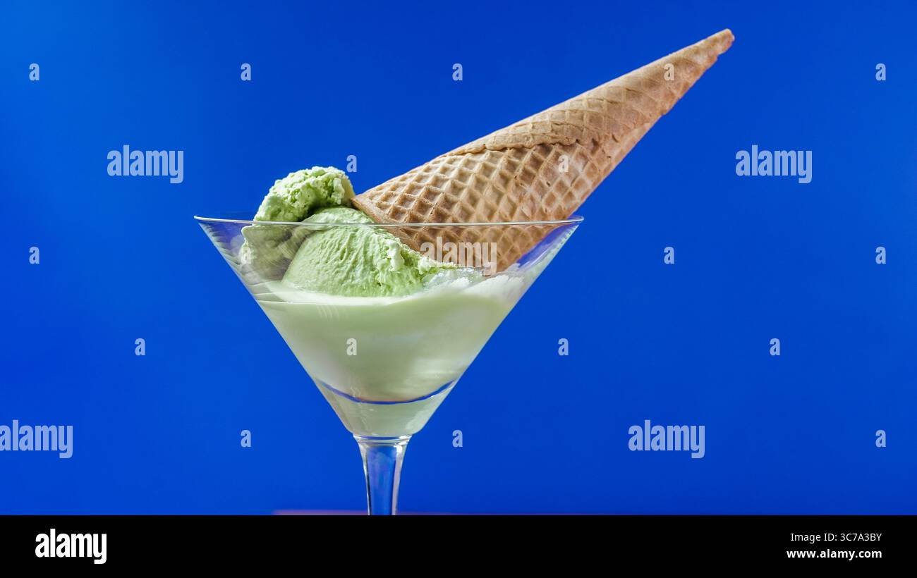 Eine erfrischende Portion Matcha-grüner Eiscreme wird elegant in eine Waffelhülle gelegt, die am Rand eines Glases ausbalanciert ist. Der leuchtend blaue Hintergrund wird noch verbessert Stockfoto