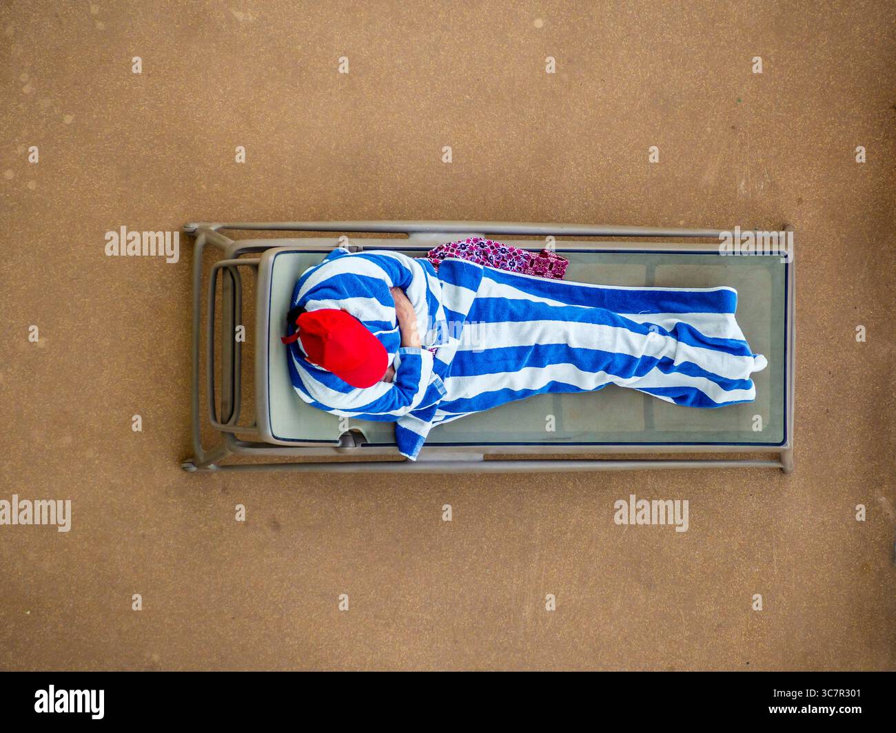 Top-down-Perspektive eines Mannes mit rotem Baseballhut, bedeckt mit einer blau-weiß gestreiften Decke, die auf einer Sonnenliege an Bord eines Kreuzfahrtschiffs liegt Stockfoto Top-down-Perspektive eines Mannes mit rotem Baseballhut, bedeckt mit einer blau-weiß gestreiften Decke, die auf einer Sonnenliege an Bord eines Kreuzfahrtschiffs liegt Stockfoto