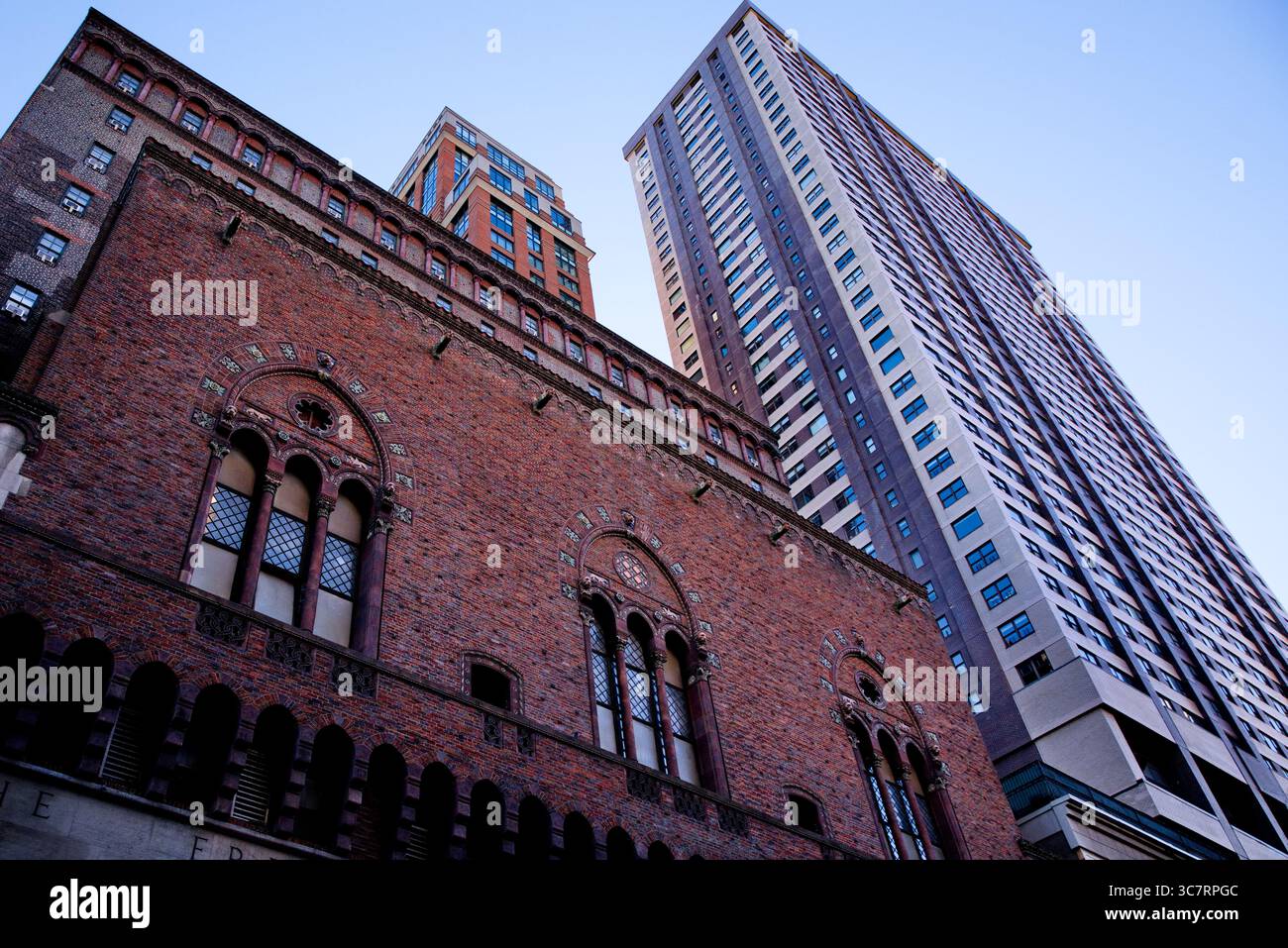 Die historische rote Backsteinfassade einer neogotischen Kirche steht unter hoch aufragenden modernen Türmen entlang einer Schlucht in Manhattan unter einem strahlend blauen Himmel. Stockfoto
