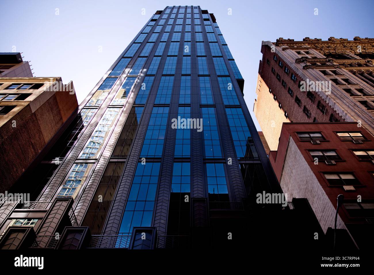 Ein eleganter blauer Glasturm erhebt sich zwischen Mauerwerk-Gebäuden in einer Manhattan-Straße. Seine reflektierende Fassade fängt die pulsierende Energie der Stadt unter einem klaren Himmel ein Stockfoto
