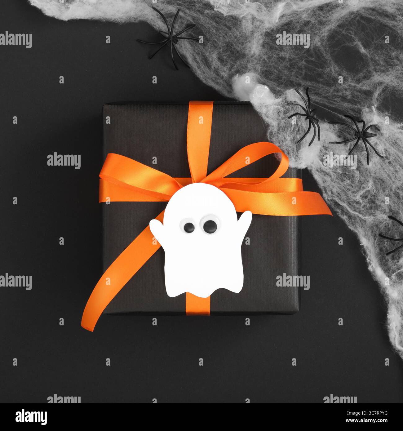 Schwarze Geschenkbox mit orangefarbenem Band und dekorativem Geist auf schwarzem Hintergrund mit Spinnennetz und Spinne. Halloween diy Geschenk Stockfoto