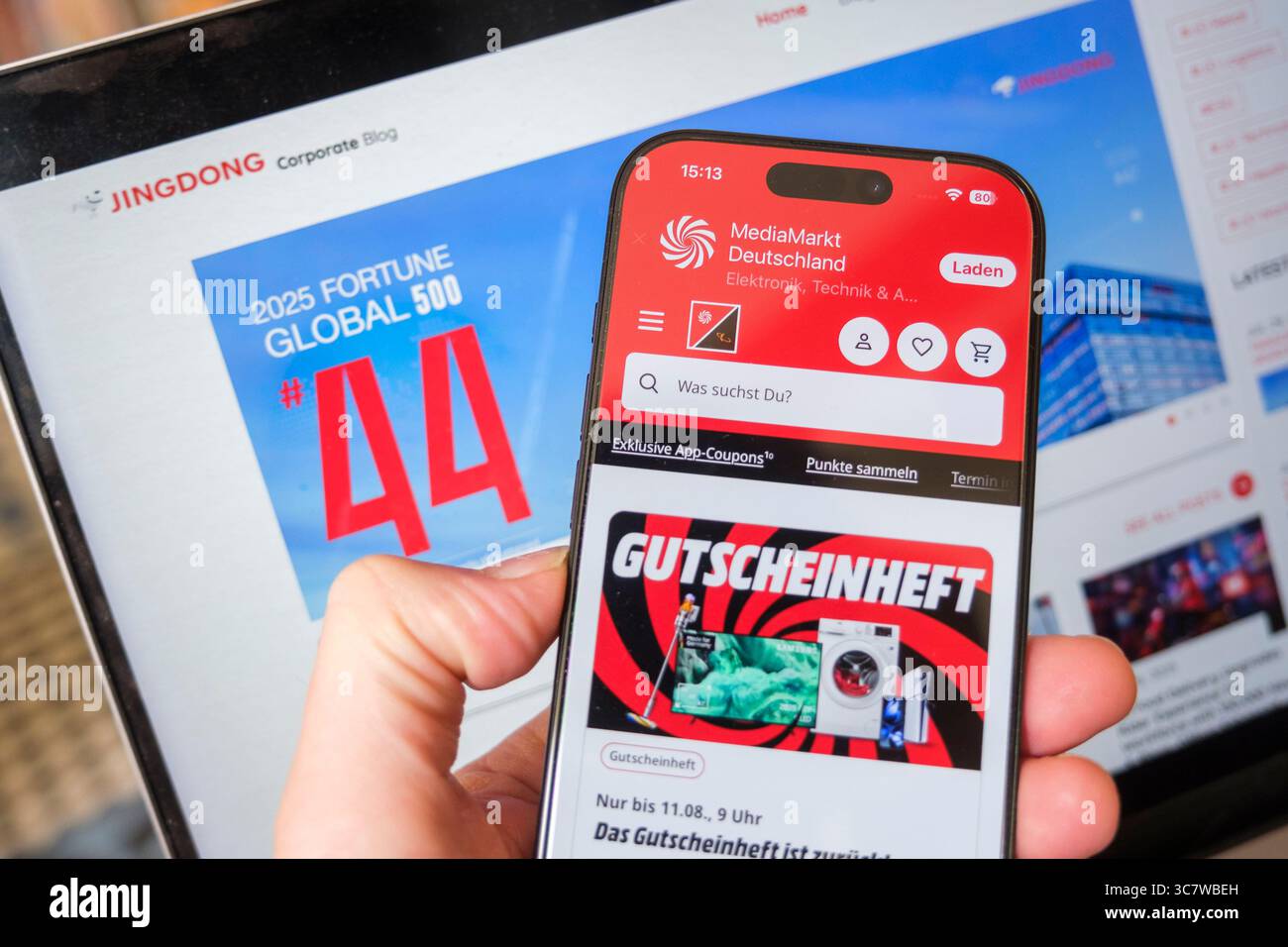 Eine Hand hält ein Smartphone mit geöffneter MediaMarkt-Website vor einem Laptop, der die Website von JD.com zeigt Stockfoto