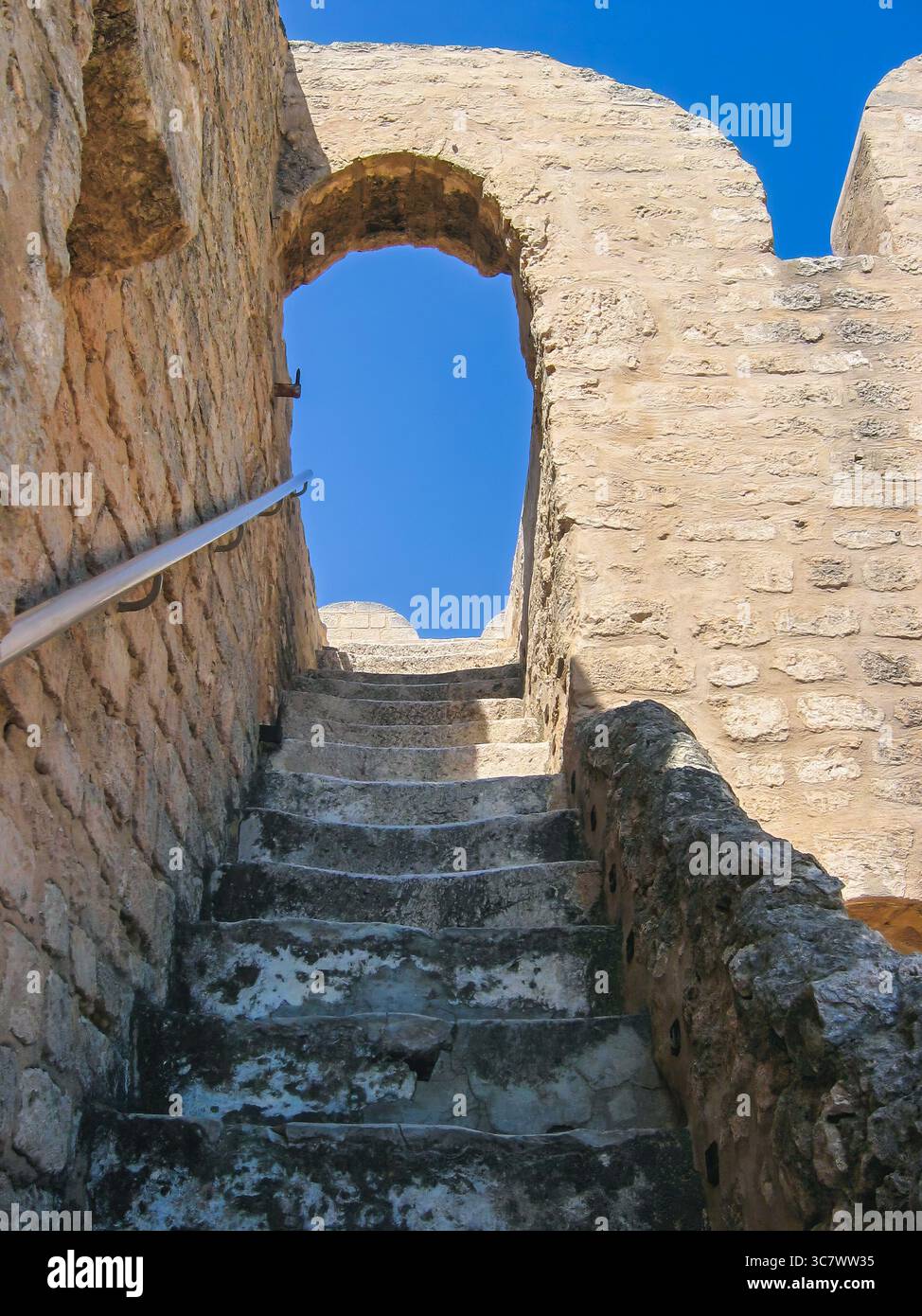 Eine antike Steintreppe führt durch eine bogenförmige Öffnung nach oben, die verwitterte Texturen und den blauen Himmel zeigt und einlädt, das Ribat of Sousse Ar zu erkunden Stockfoto
