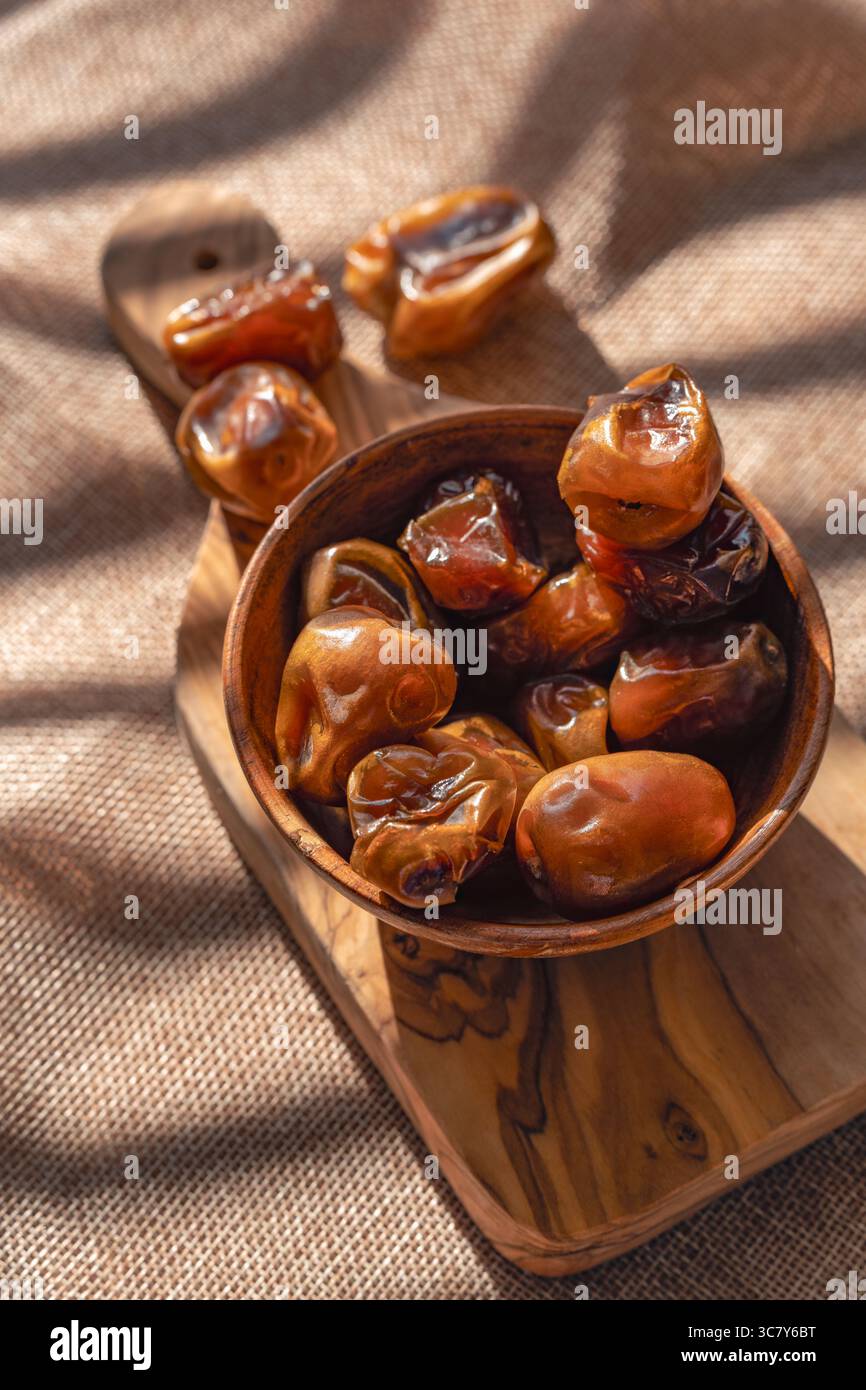 Eine braune Holzschale voller getrockneter Datteln, die auf einem Schneidebrett aus natürlichem Holz platziert wird, mit Sonnenlicht, das künstlerische Schatten schafft und Text hervorhebt Stockfoto