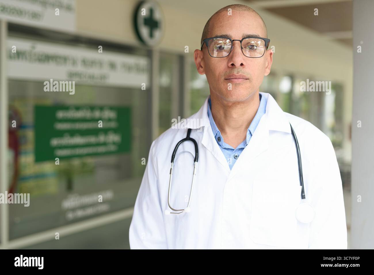 Reifer männlicher Arzt trägt Labormantel und Brille, der selbstbewusst vor einer kleinen Gesundheitsklinik steht. Medizinisches Fachkonzept für den Außenbereich. Stockfoto