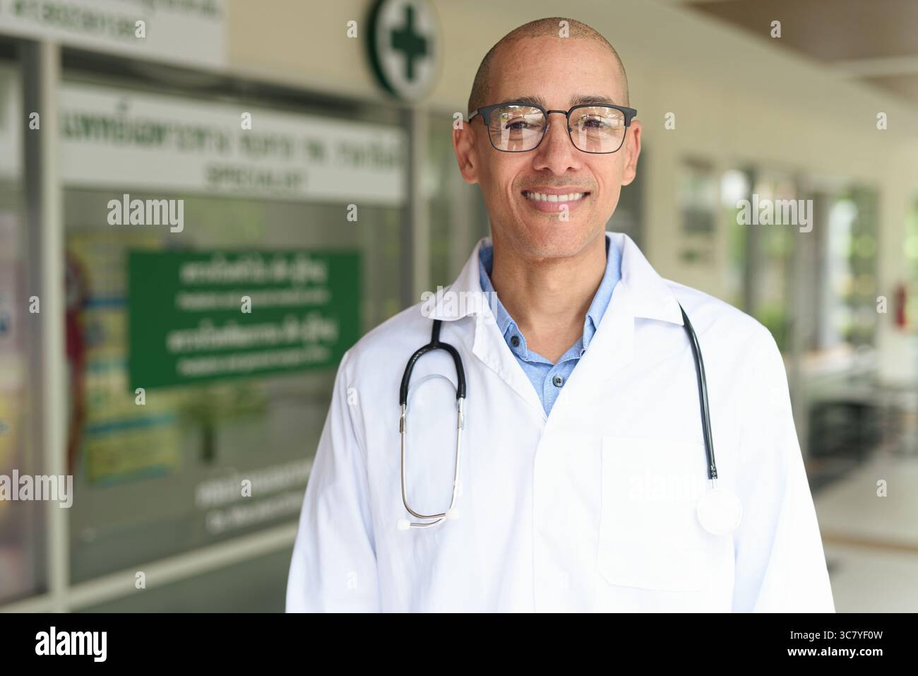 Reifer männlicher Arzt trägt Labormantel und Brille, der selbstbewusst vor einer kleinen Gesundheitsklinik steht. Medizinisches Fachkonzept für den Außenbereich. Stockfoto