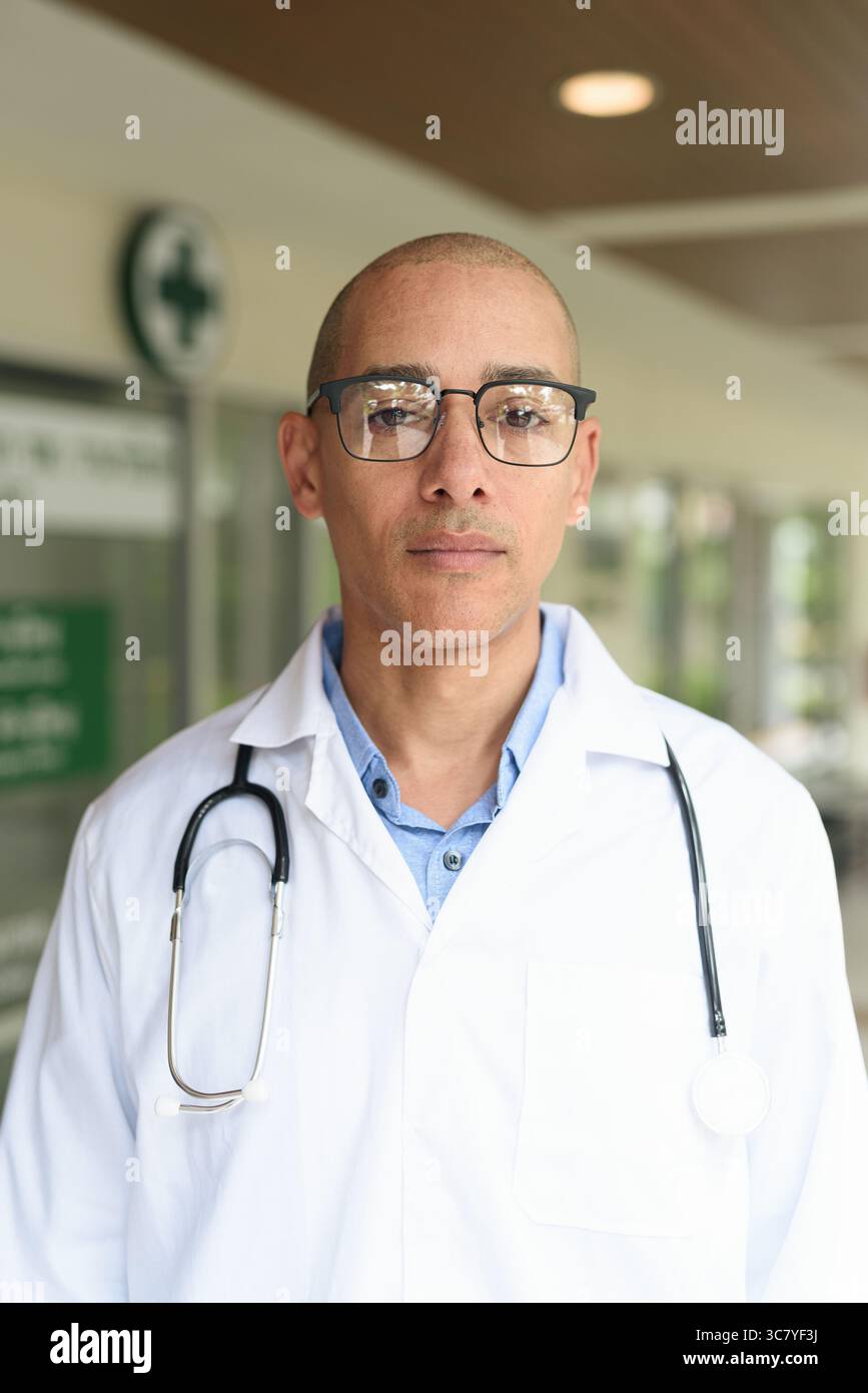 Reifer männlicher Arzt trägt Labormantel und Brille, der selbstbewusst vor einer kleinen Gesundheitsklinik steht. Medizinisches Fachkonzept für den Außenbereich. Stockfoto